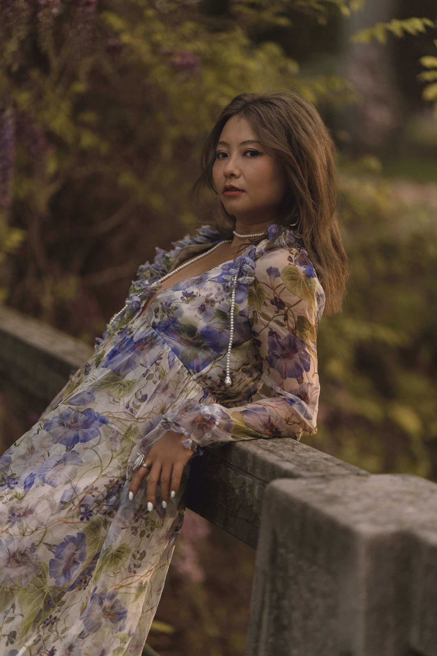 Woman in blue floral dress leaning on stone wall in garden setting.