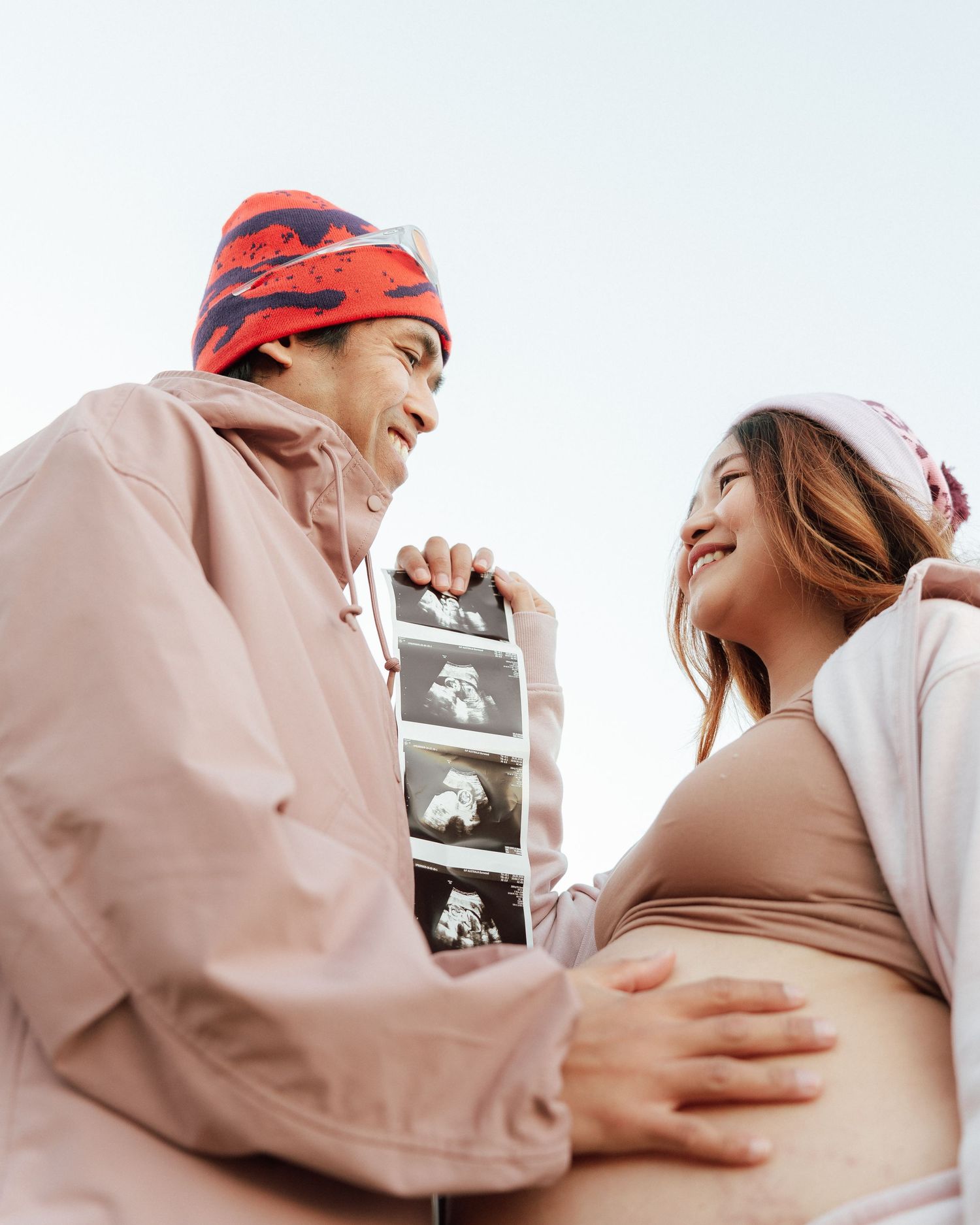 Joyful expecting couple in matching pink outfits share ultrasound photos while partner touches pregnant belly.