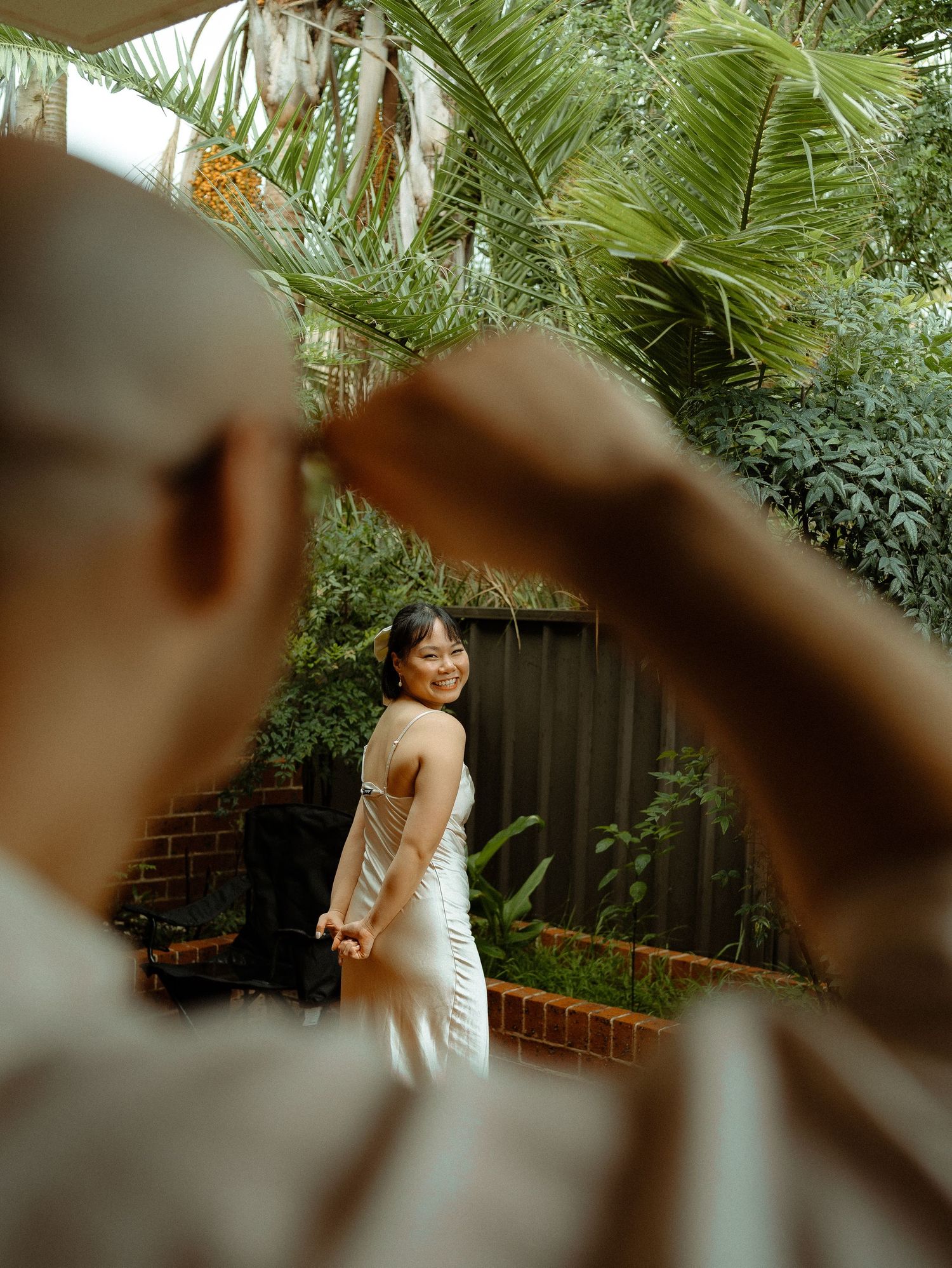 Couple holding hands in a lush garden, woman in white dress smiling over her shoulder.