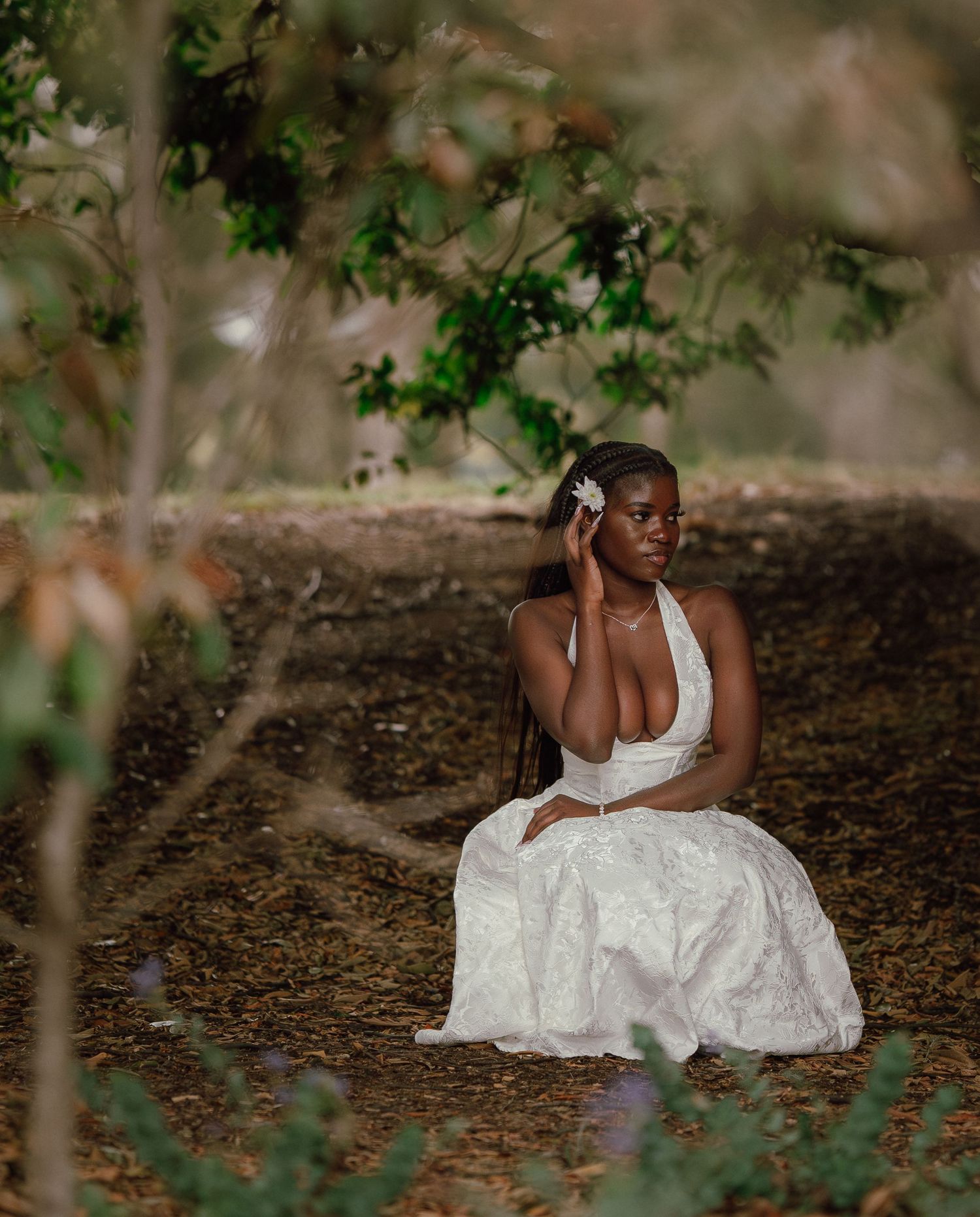 A young Black woman in a white halter dress sits on the ground in a wooded area with a flower in her hair.