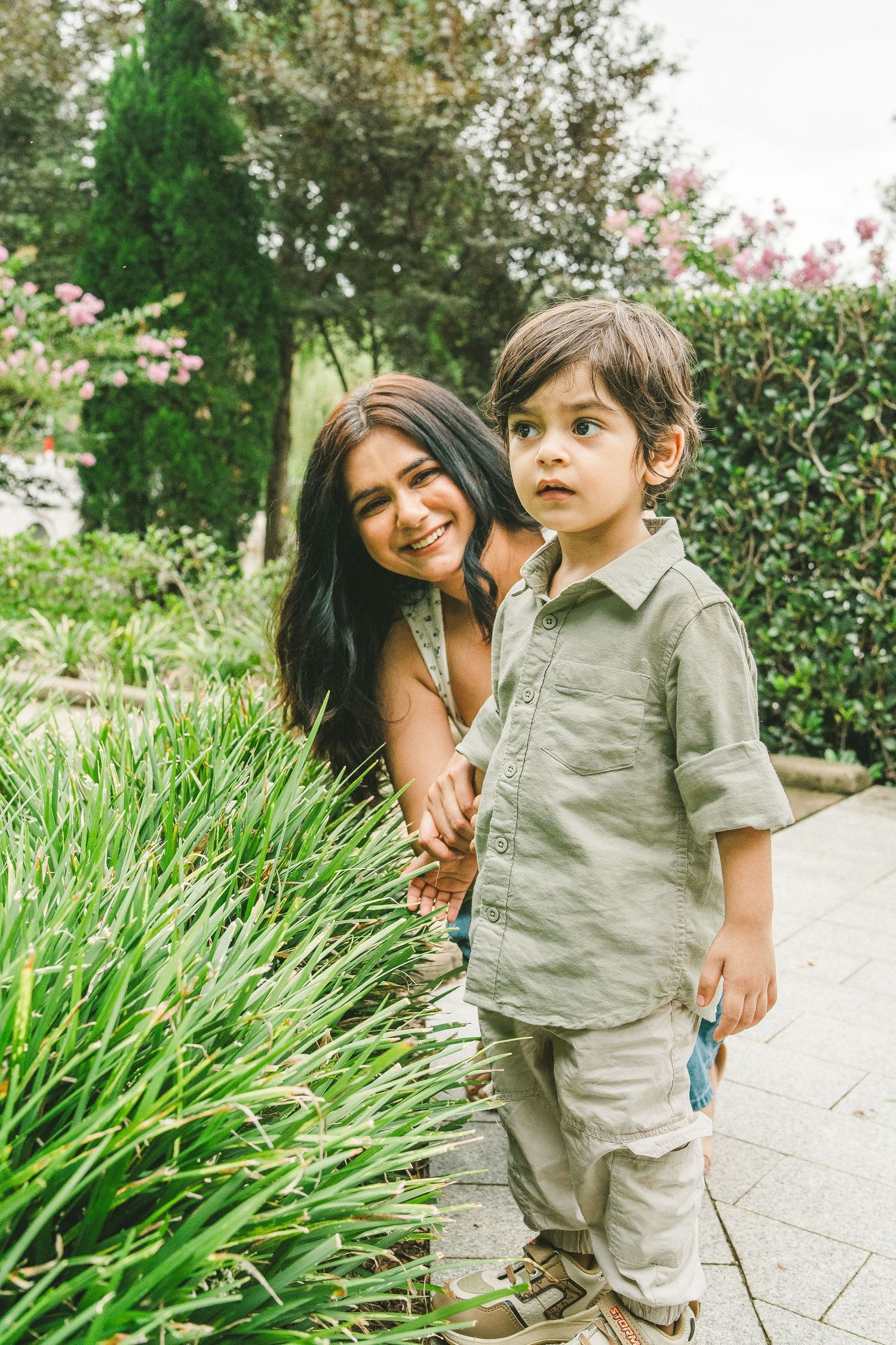 Smiling mother crouching beside curious toddler son near lush green garden plants on a pathway.