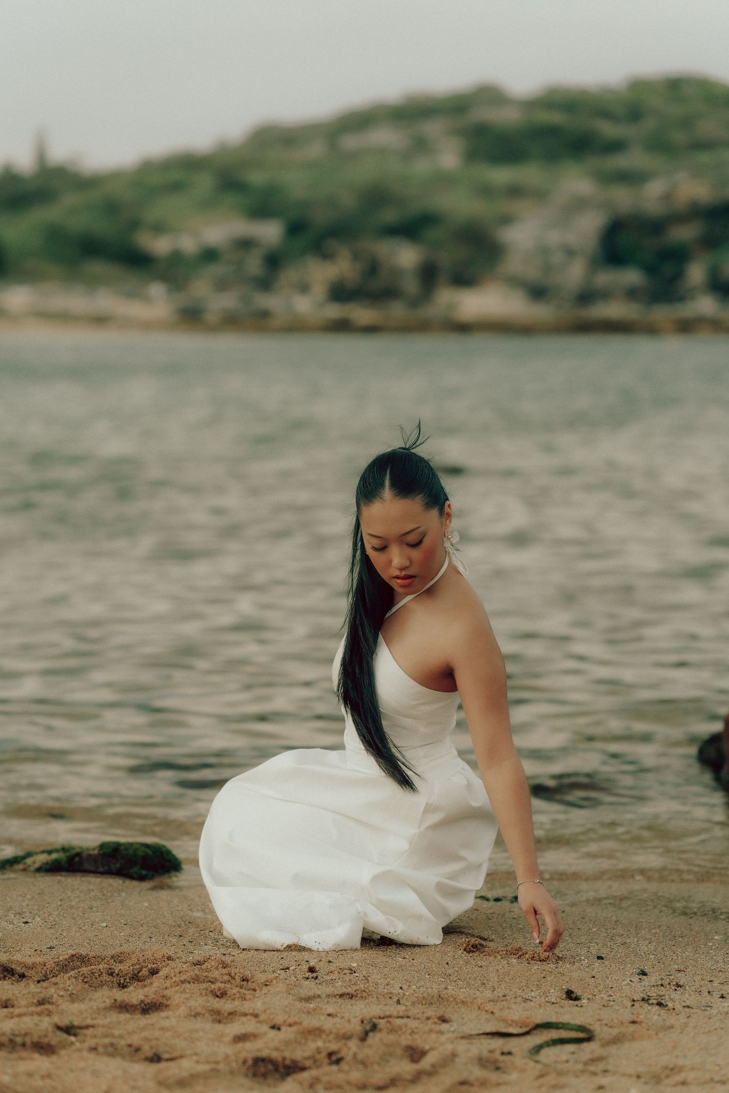 Young Asian woman in white dress crouching on sandy beach near calm water's edge.
