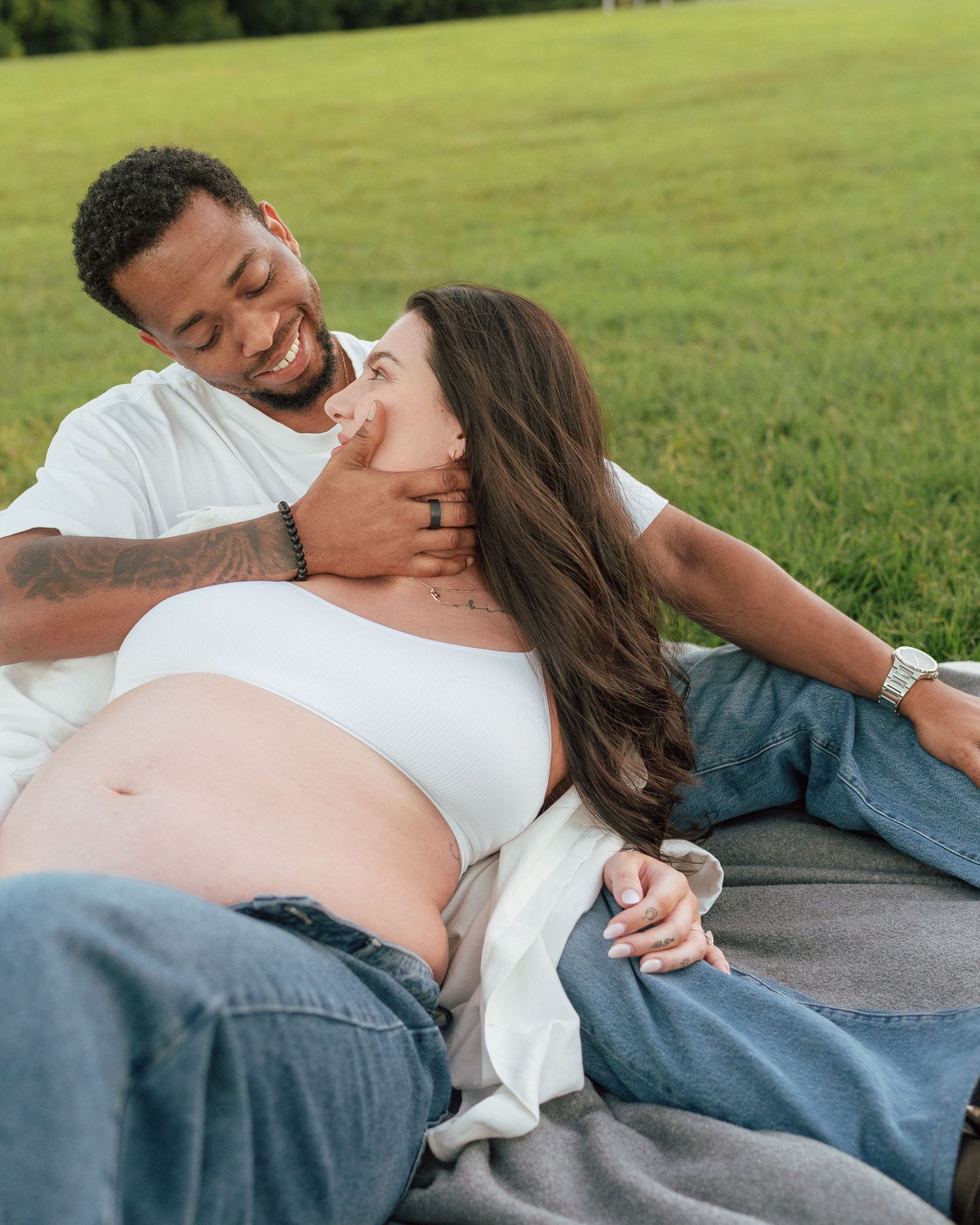 Expectant couple relaxing on a blanket in a green park, man tenderly touching pregnant womans face.