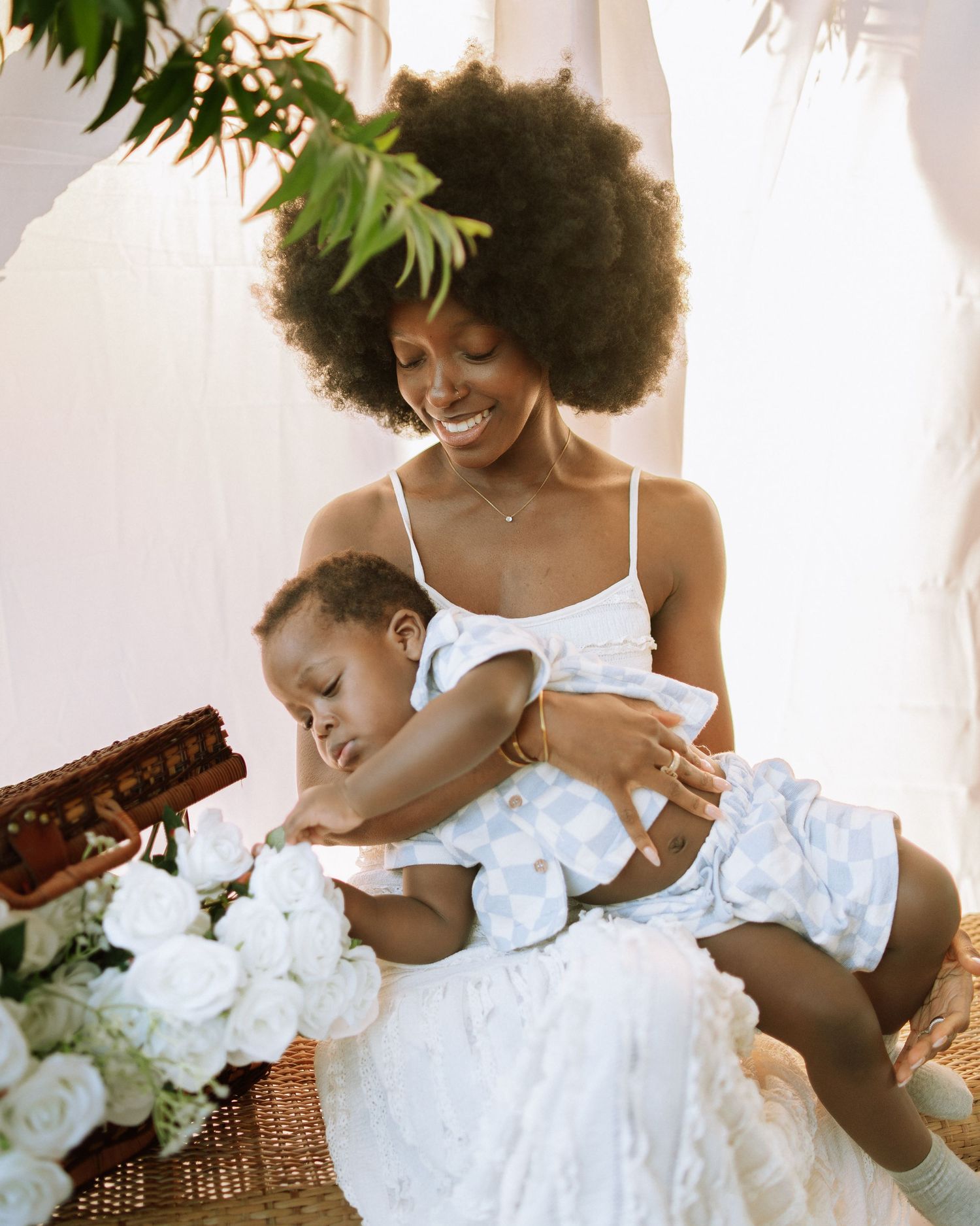 A smiling mother with an afro holds her laughing baby surrounded by white roses in a bright outdoor setting.
