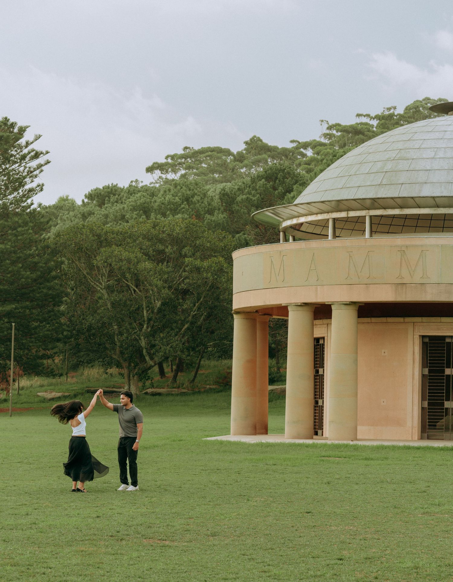 A couple dances and twirls together on a green lawn beside a domed circular building with columns.