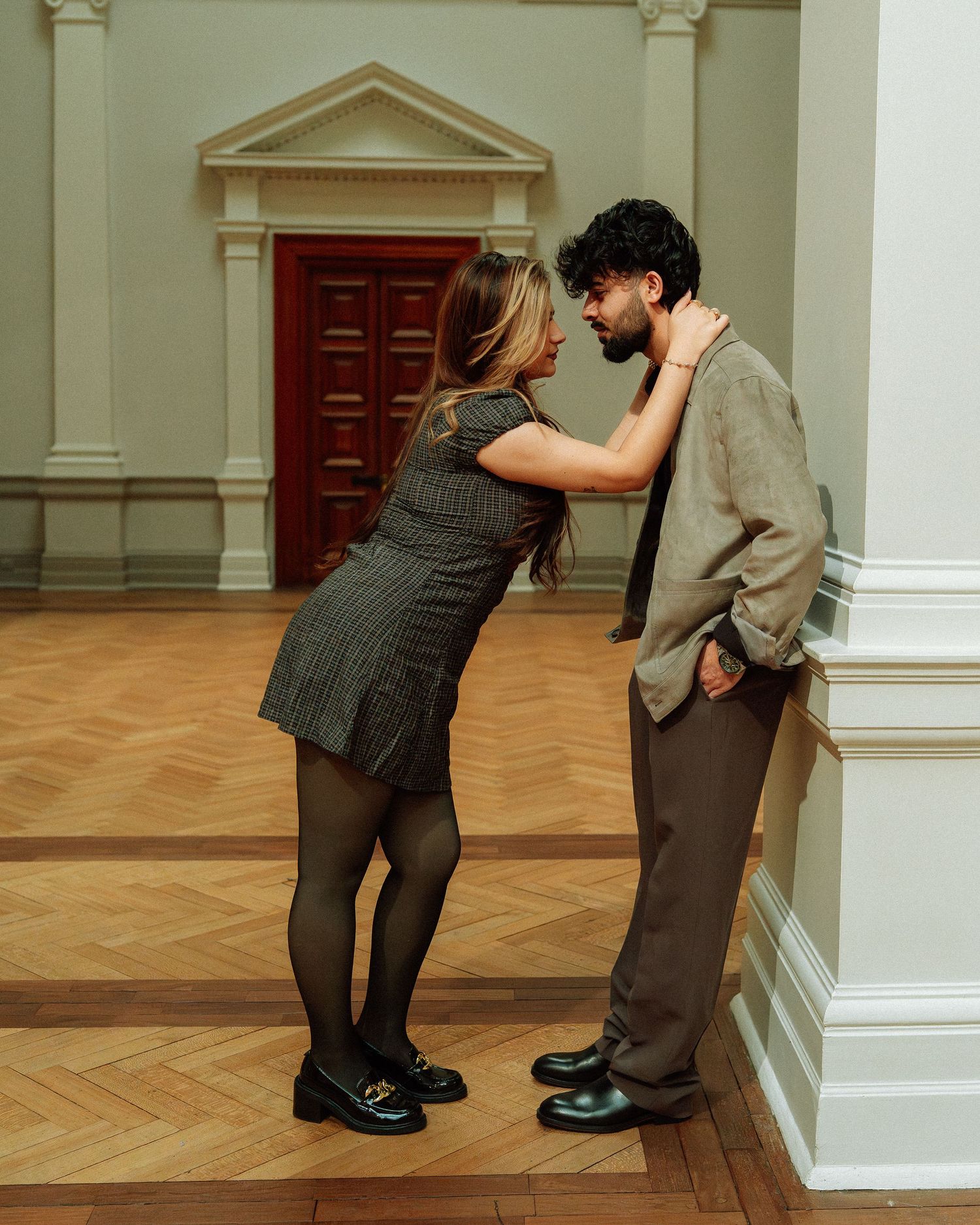 Couple sharing an intimate moment leaning against a white column in an elegant museum hall.