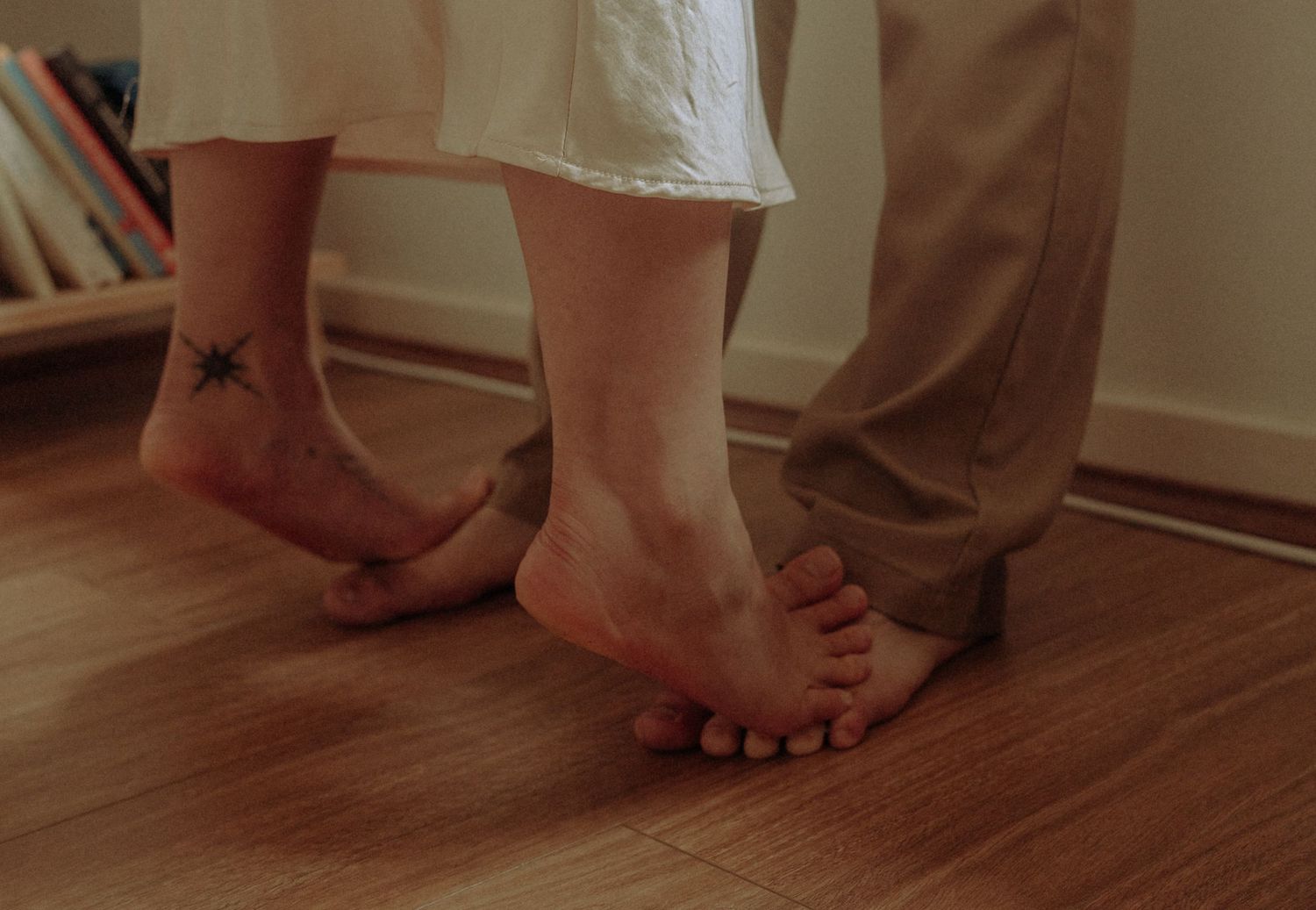 Close-up of two pairs of bare feet on hardwood floor, one ankle adorned with a star tattoo.