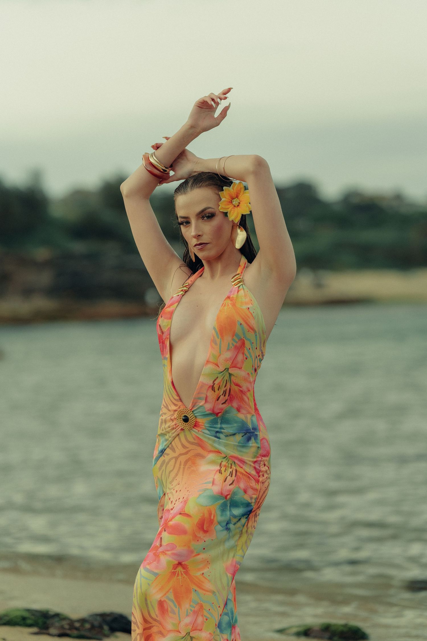 Woman in colorful tropical halter dress poses on beach with arms raised overhead and yellow flower in hair.