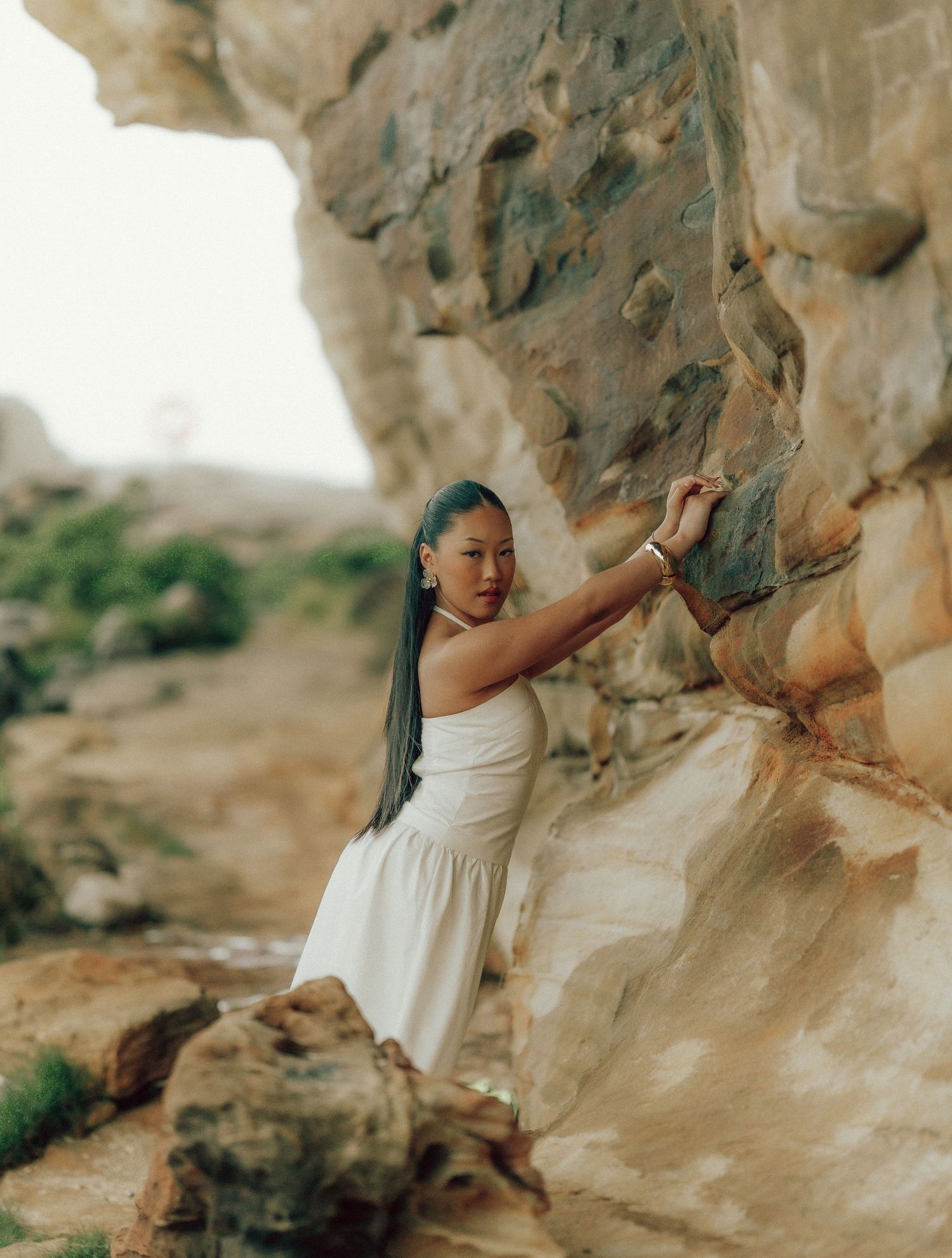 Asian woman in white dress reaching up to touch textured sandstone rock wall in natural setting.