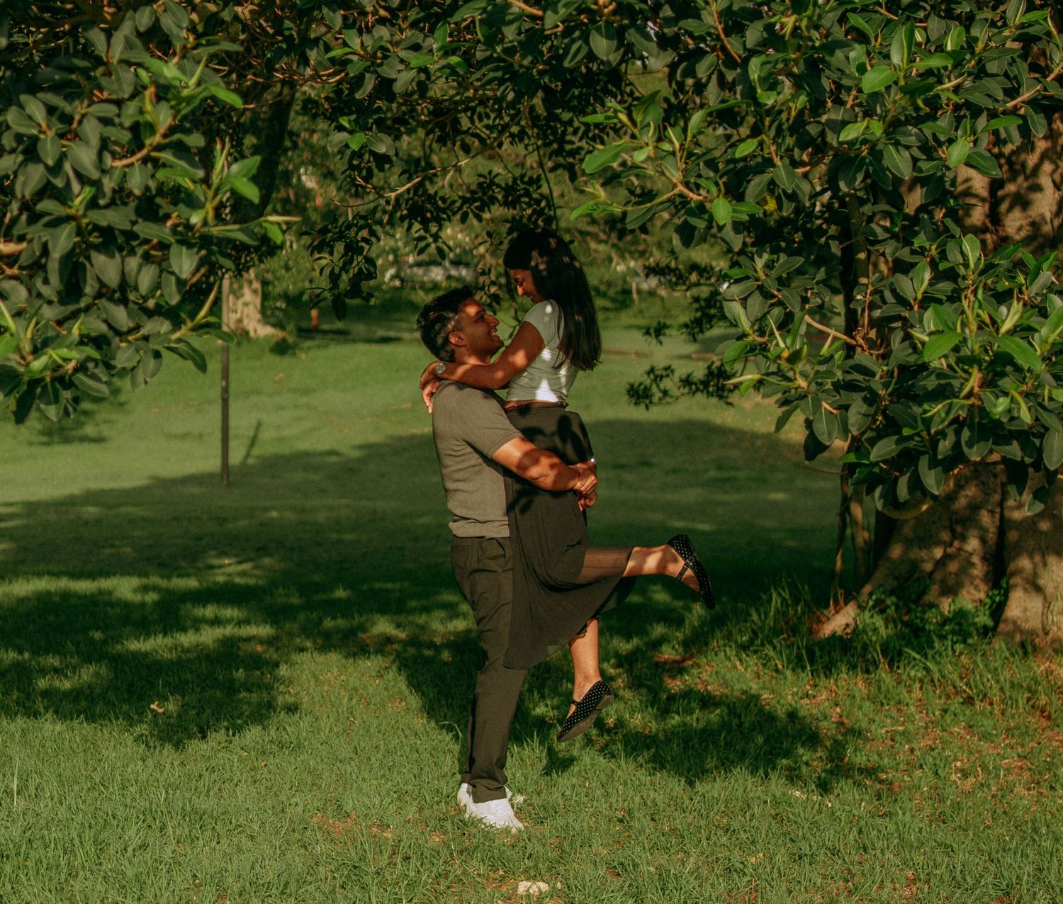Couple sharing a romantic embrace under lush green trees in a sunlit park.