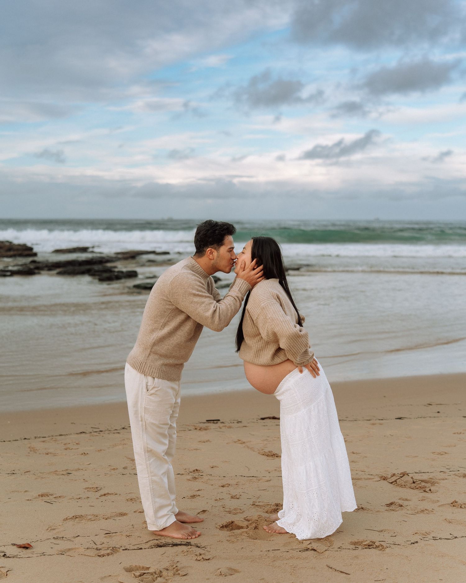 Expecting couple shares a romantic kiss on sandy beach with ocean waves, wearing neutral tones for maternity photos.