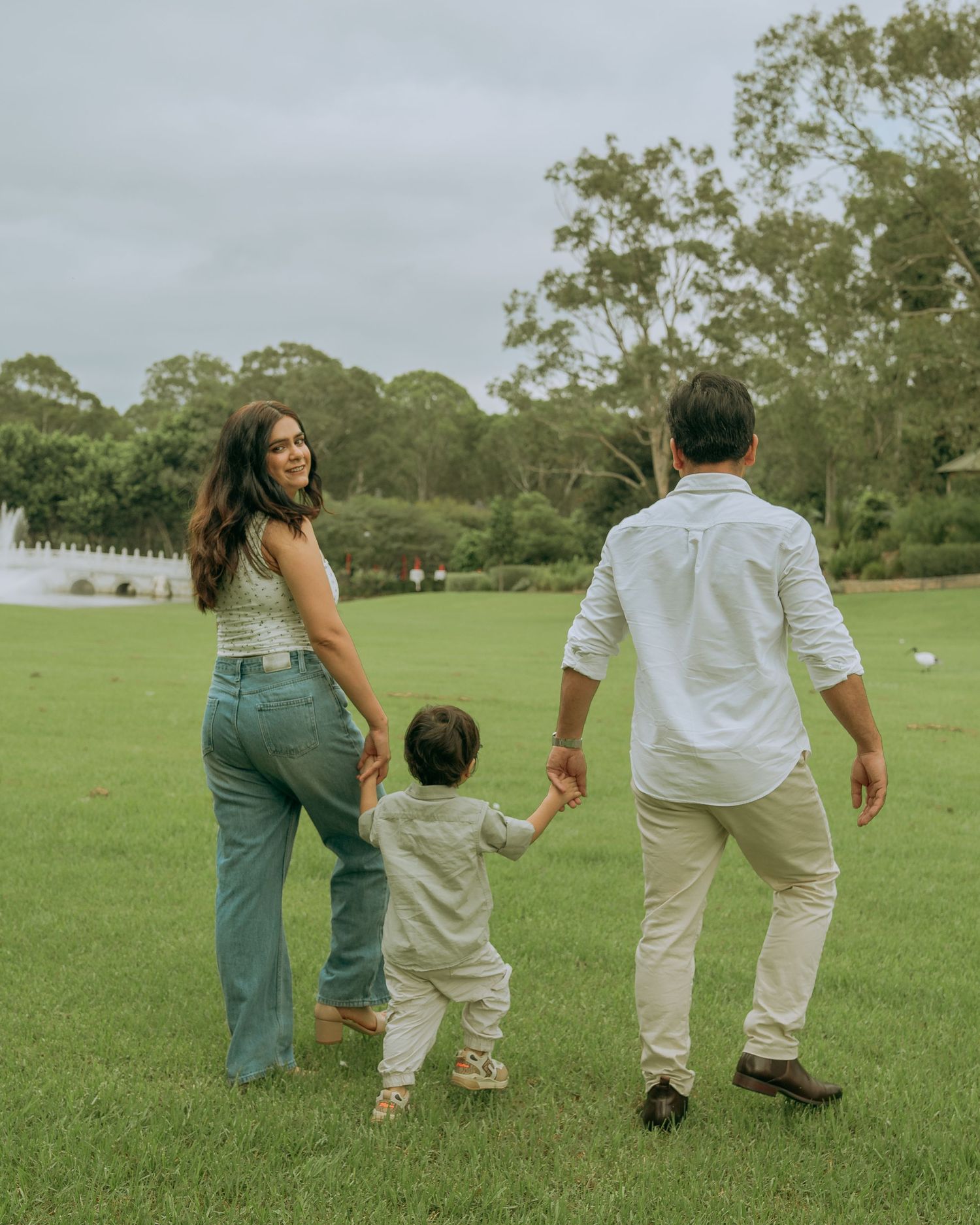 Young family holding hands walking together across a lush green park lawn.