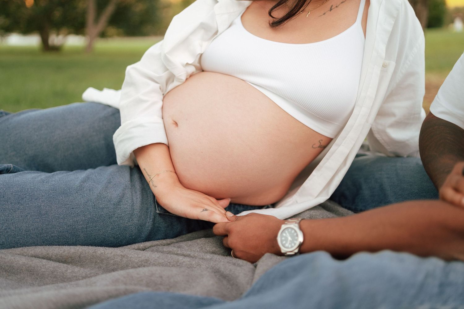 Close-up of pregnant womans bare baby bump with partners hand resting gently on her stomach outdoors.