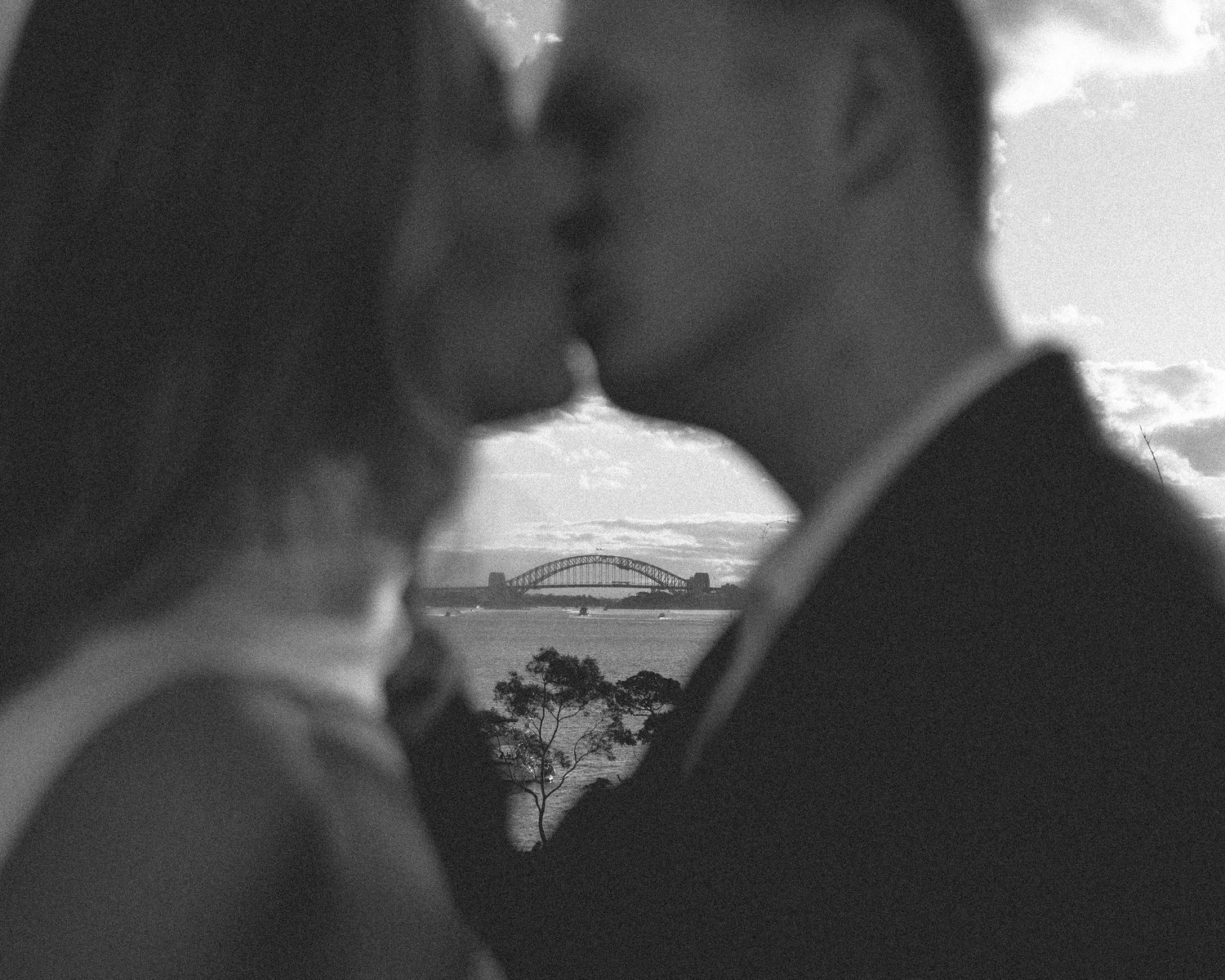 Black and white photo of newlyweds kissing with Sydney Harbour Bridge silhouetted in background.