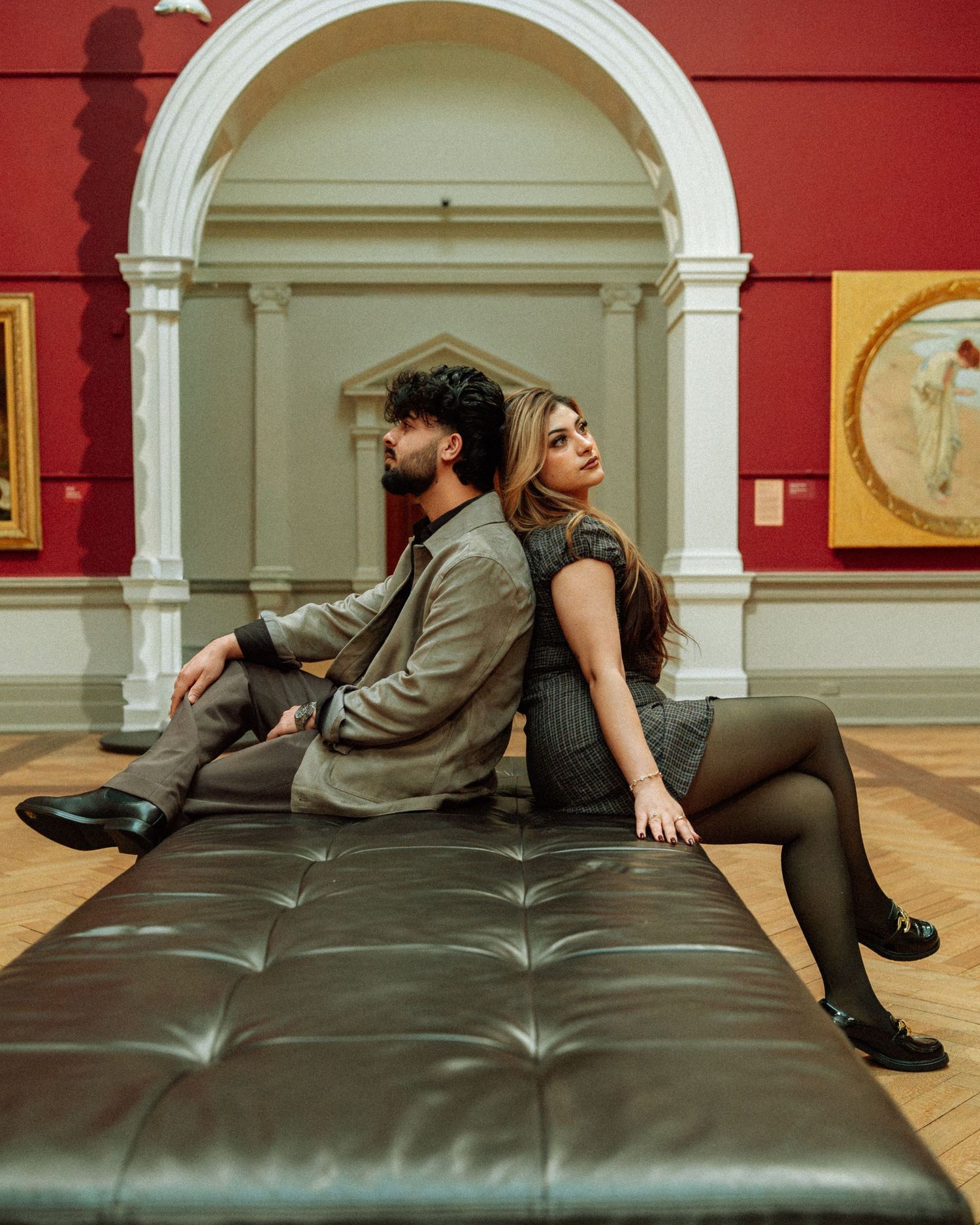 Couple sitting back-to-back on a leather bench inside an elegant art museum with red walls.