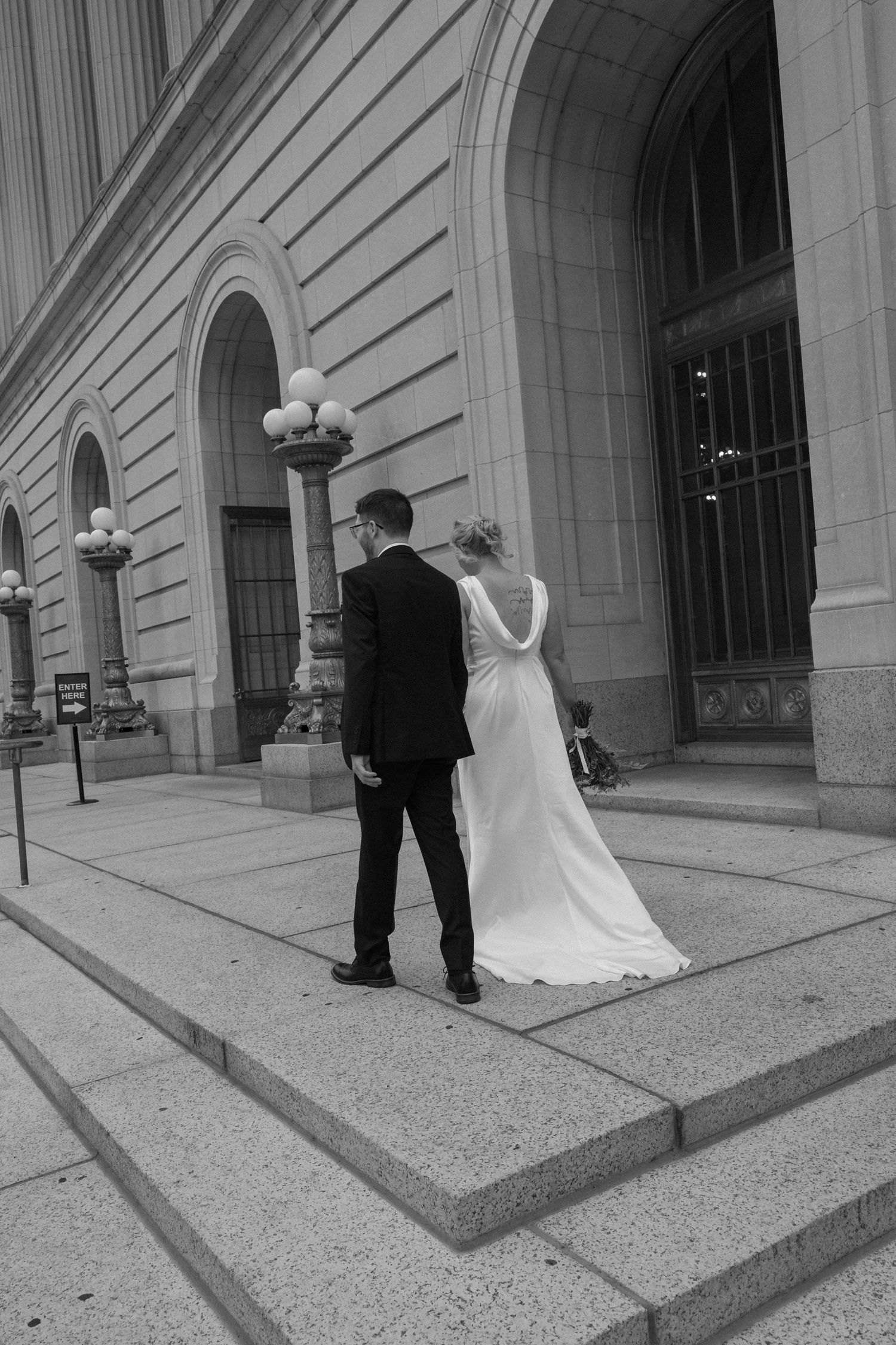 Black and white photo of bride and groom walking up steps of elegant stone building.