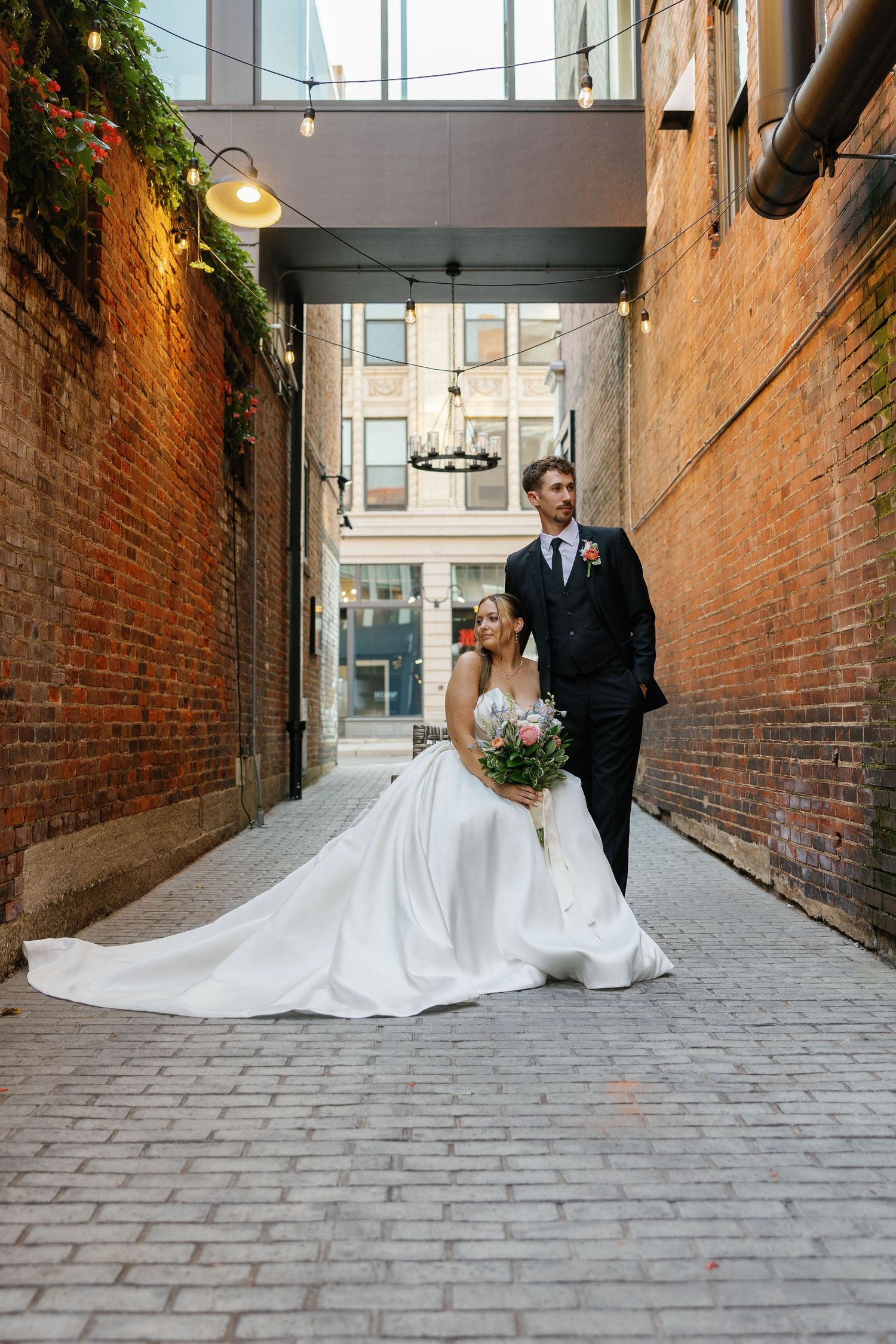 Bride in a stunning white ballgown sits on cobblestones as groom stands beside her in a brick alleyway with string lights.