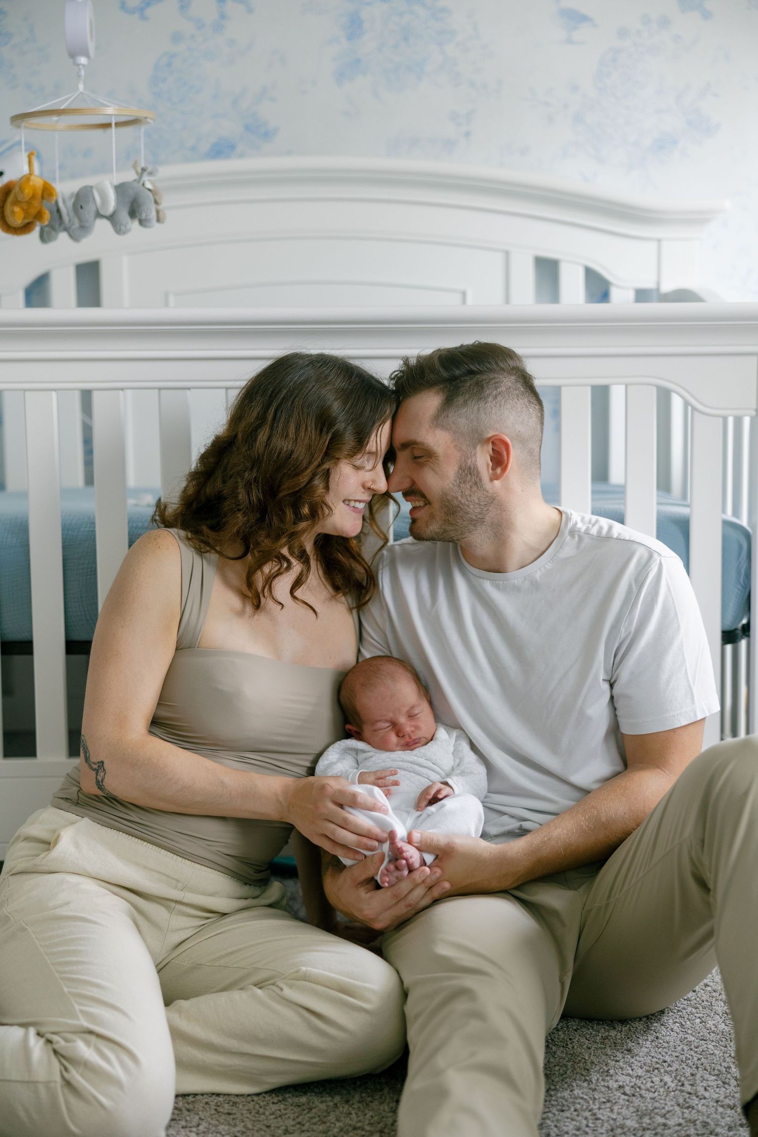 Happy couple sitting in front of white crib in nursery, tenderly holding their sleeping newborn baby.