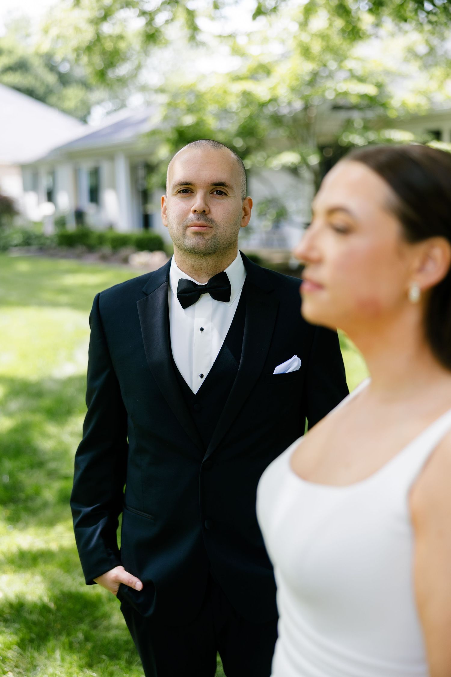 Groom in black tuxedo with bow tie gazes at bride in white dress during outdoor wedding ceremony.