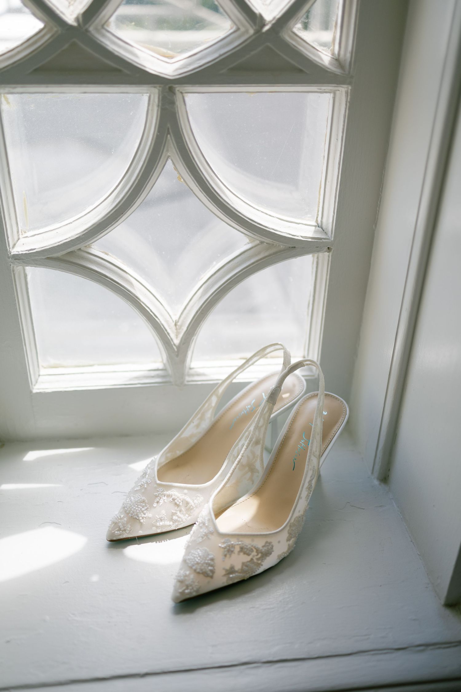 Lace slingback bridal heels displayed on a windowsill with decorative leaded glass window.