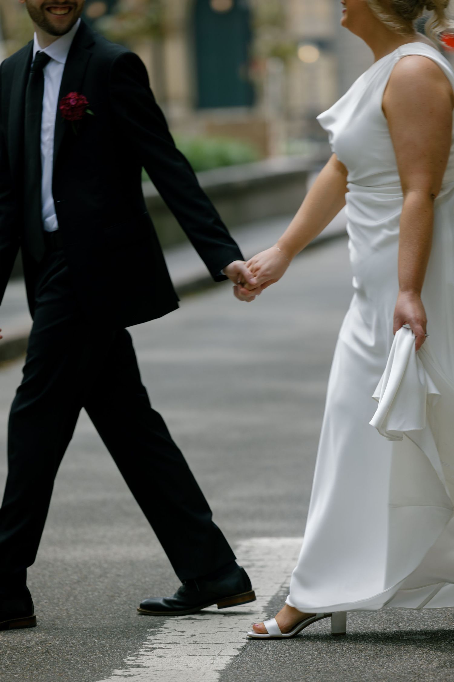 Bride in white satin gown and groom in black suit holding hands while walking street.