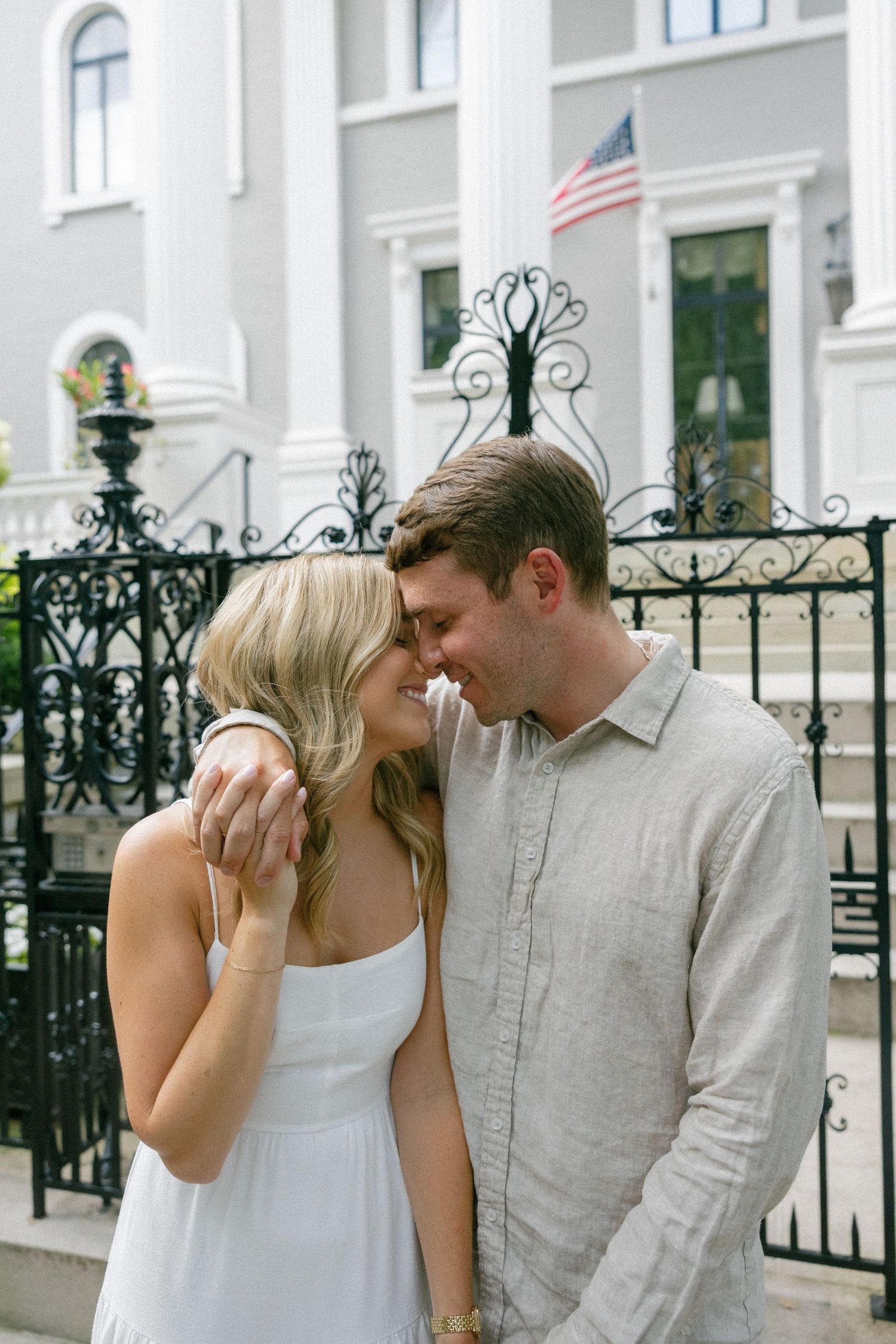 Couple sharing a romantic moment in front of an ornate iron fence and white building with American flag.