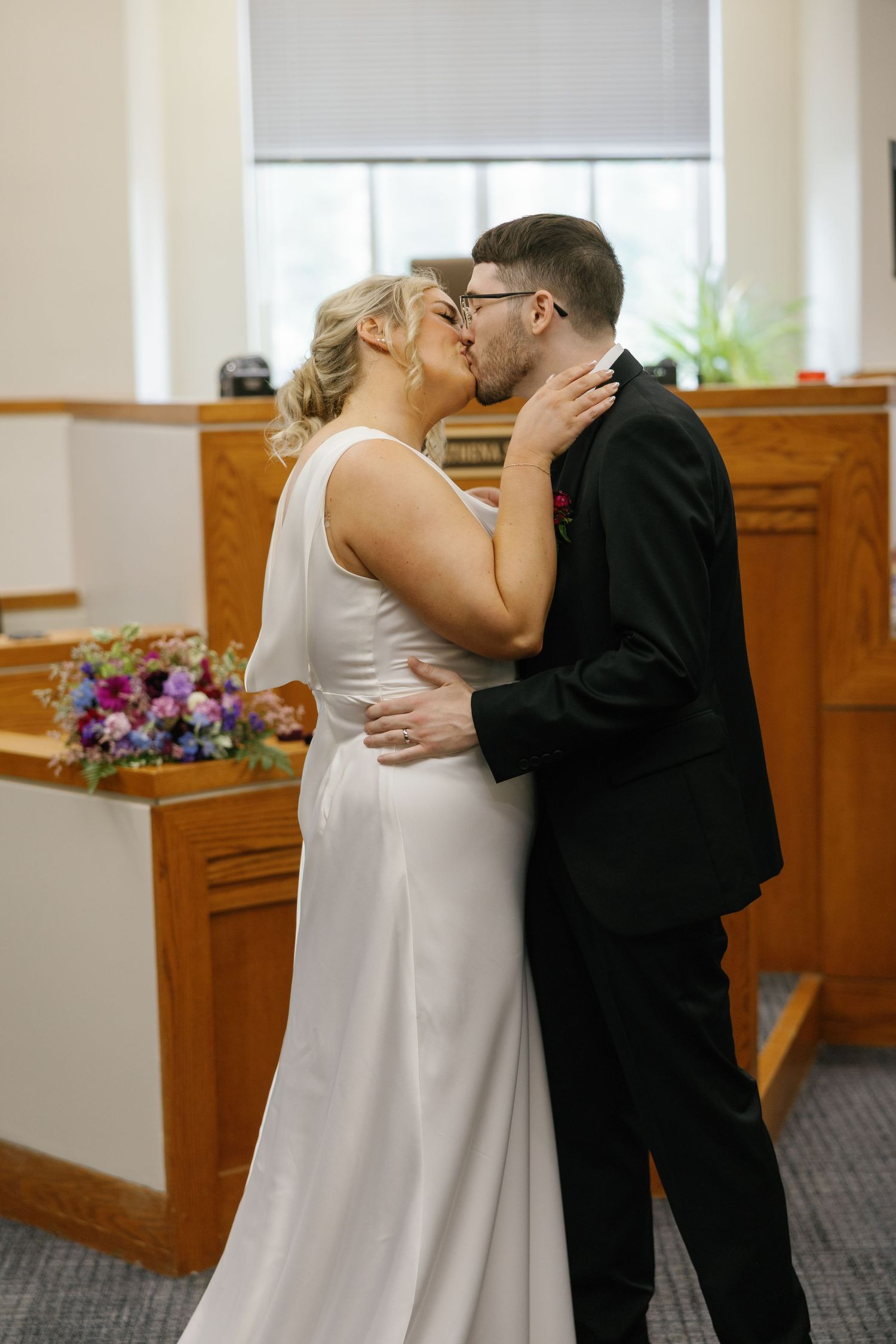 Bride and groom sharing first kiss at courthouse wedding ceremony with floral arrangement.