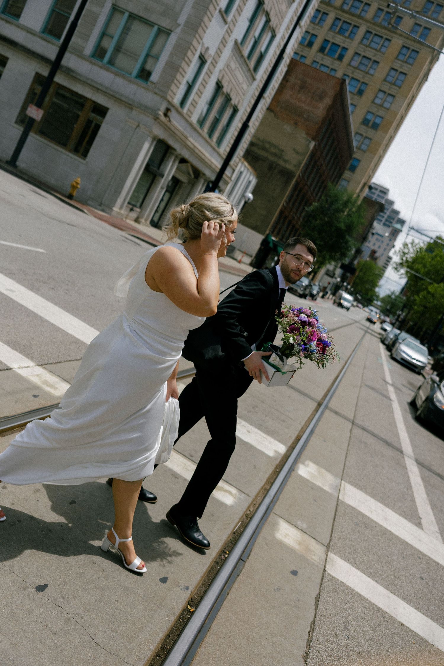 Bride and groom in wedding attire crossing a city street holding a colorful bouquet.