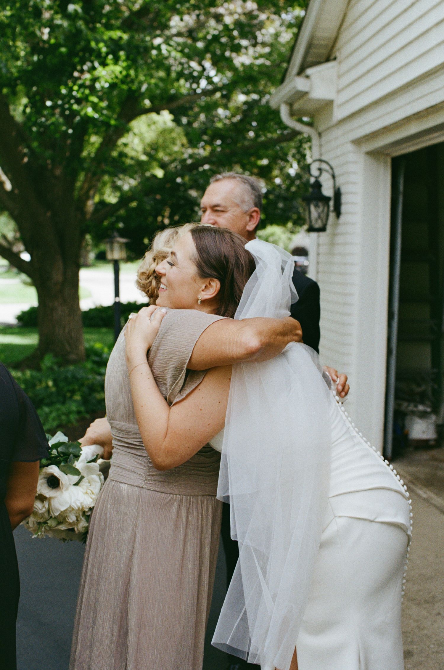 Bride in white wedding gown with veil shares warm embrace with bridesmaid at outdoor wedding.