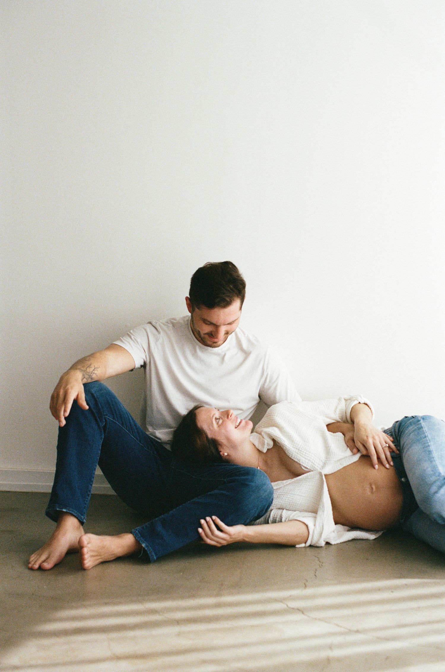 Expecting couple relaxing on floor, pregnant woman leaning back laughing joyfully while partner gazes at her.