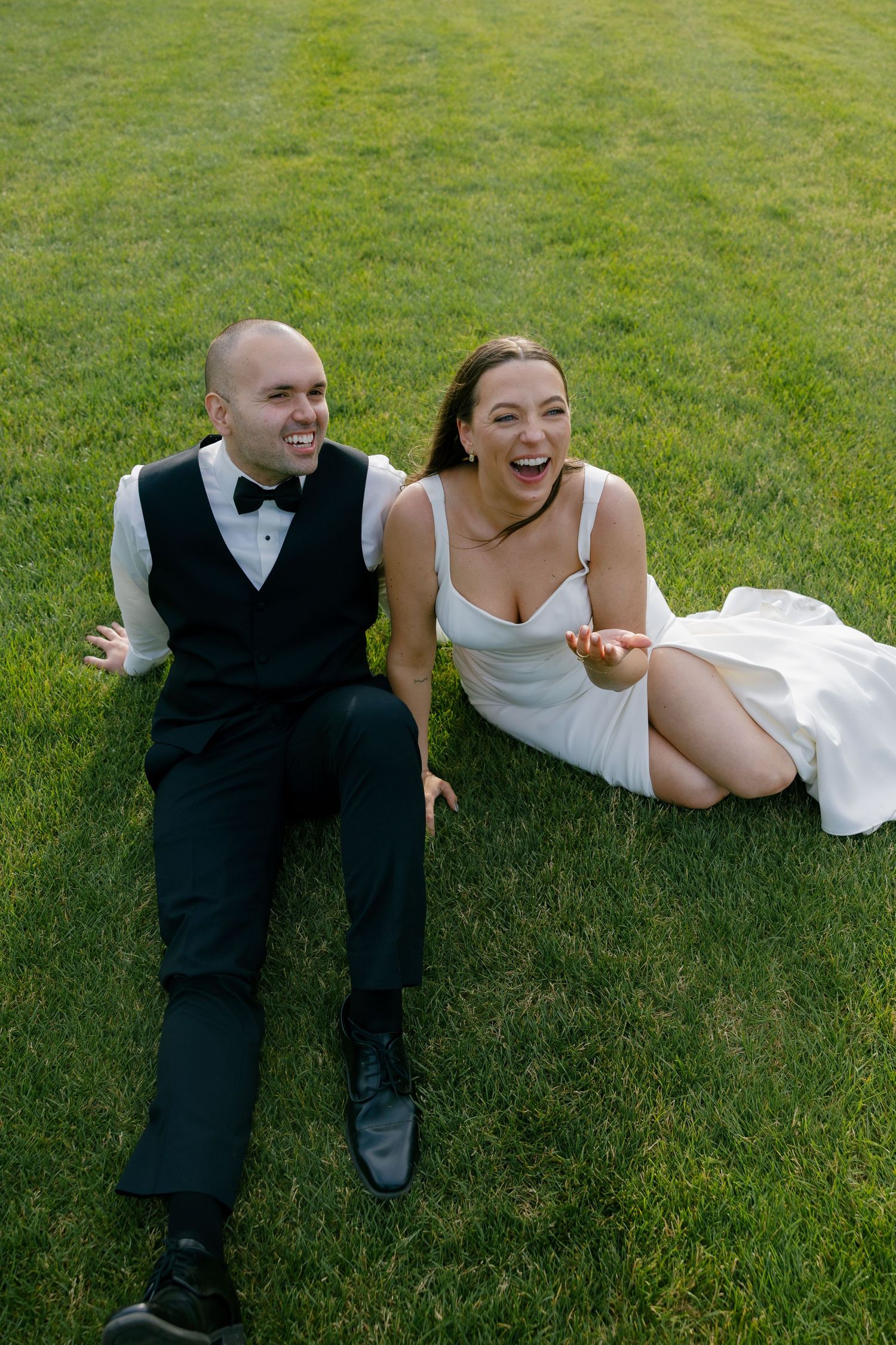 Laughing bride and groom sitting together on lush green grass during wedding celebration.