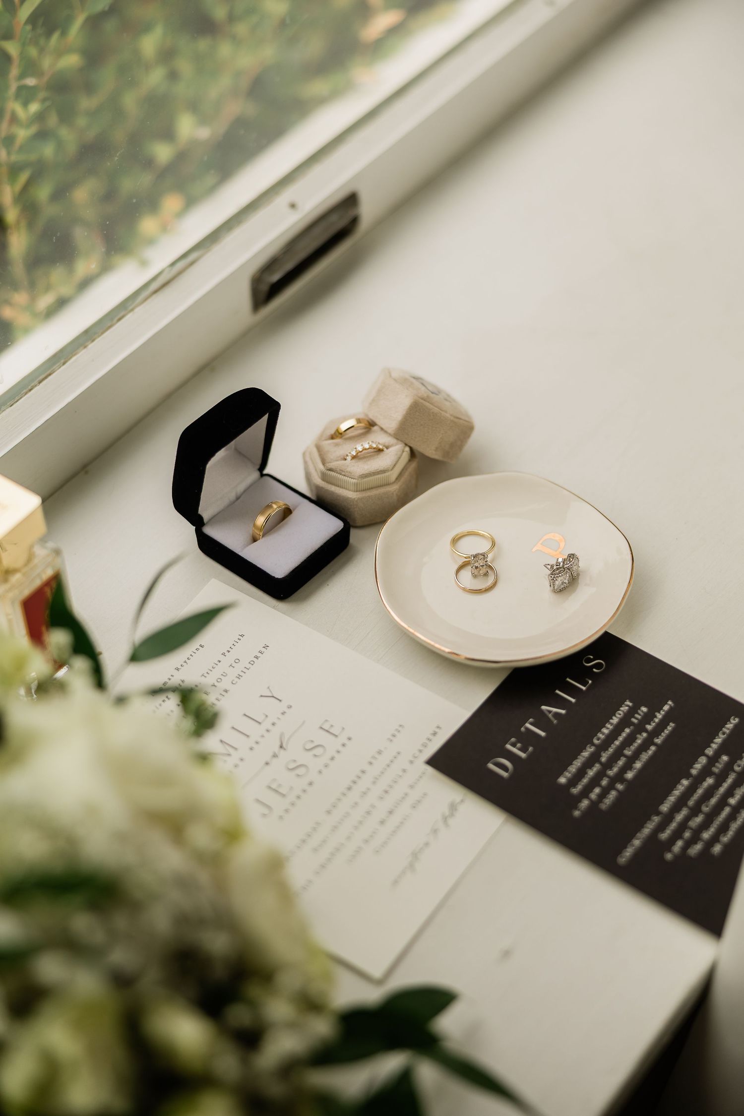 Wedding flat lay with gold rings, velvet ring boxes, invitations, and greenery on windowsill.