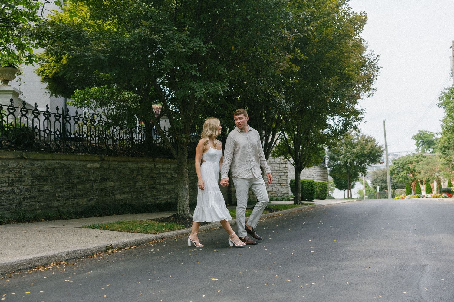 Couple walking hand in hand down a tree-lined street, dressed in elegant white and grey outfits.