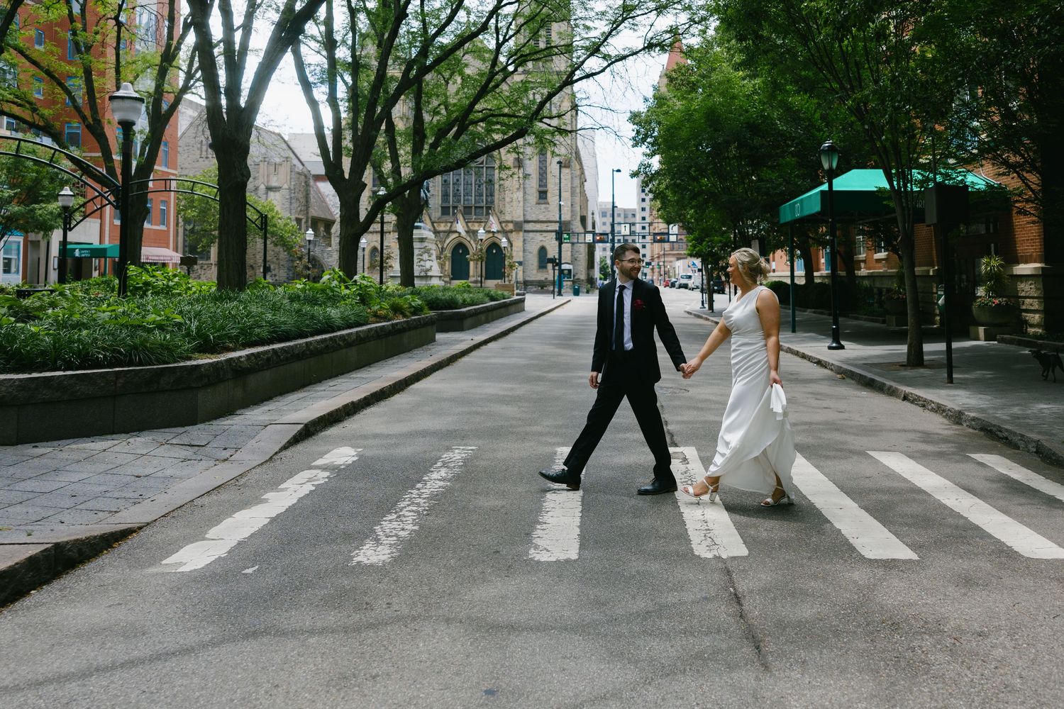 Bride and groom holding hands crossing zebra crosswalk in urban city street setting.