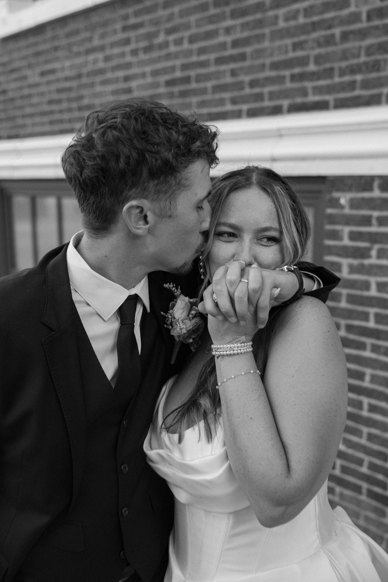 Black and white photo of groom kissing bride on the cheek as she smiles and covers her mouth with clasped hands.
