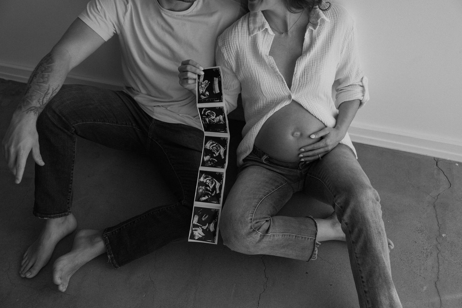 Black and white photo of couple sitting holding ultrasound strip beside pregnant belly.