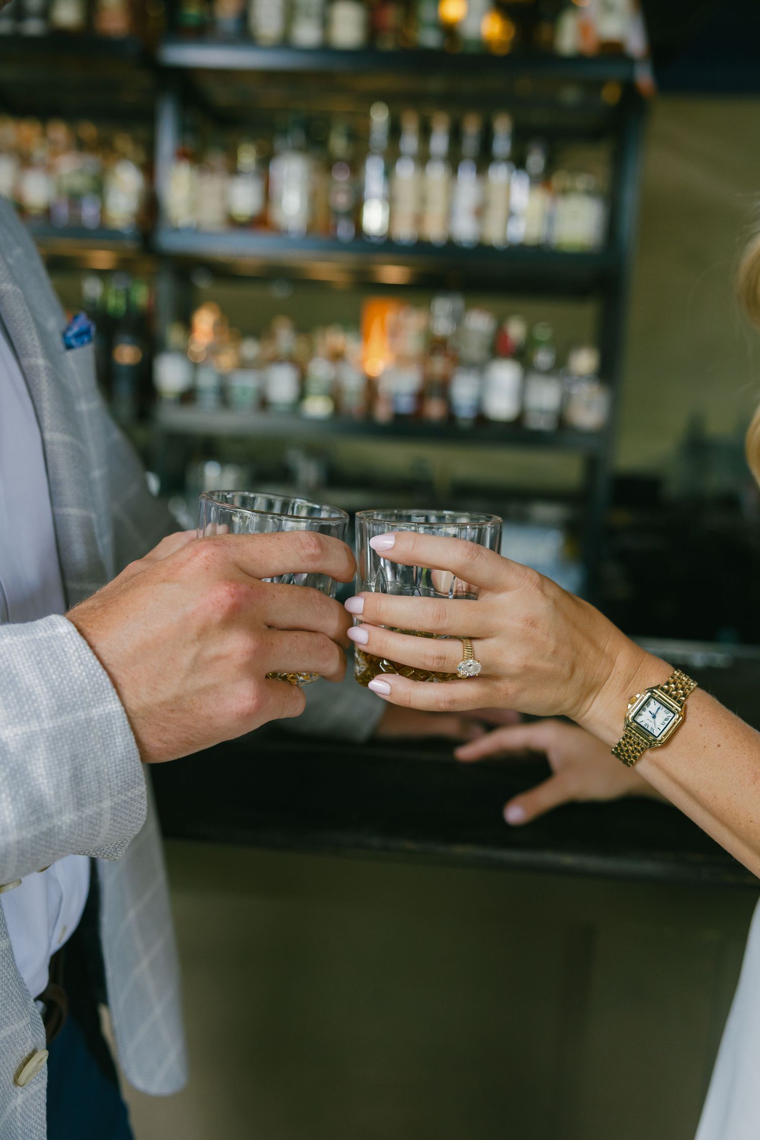 Close-up of couple toasting with whiskey glasses at a bar, woman wearing diamond engagement ring.