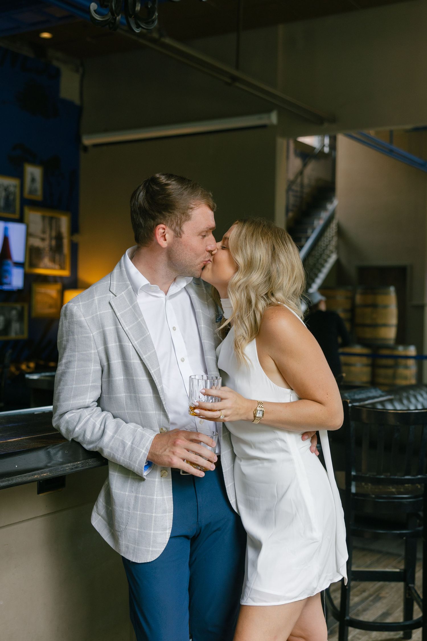 Engaged couple sharing a kiss at a bar, man in plaid blazer and woman in white mini dress.