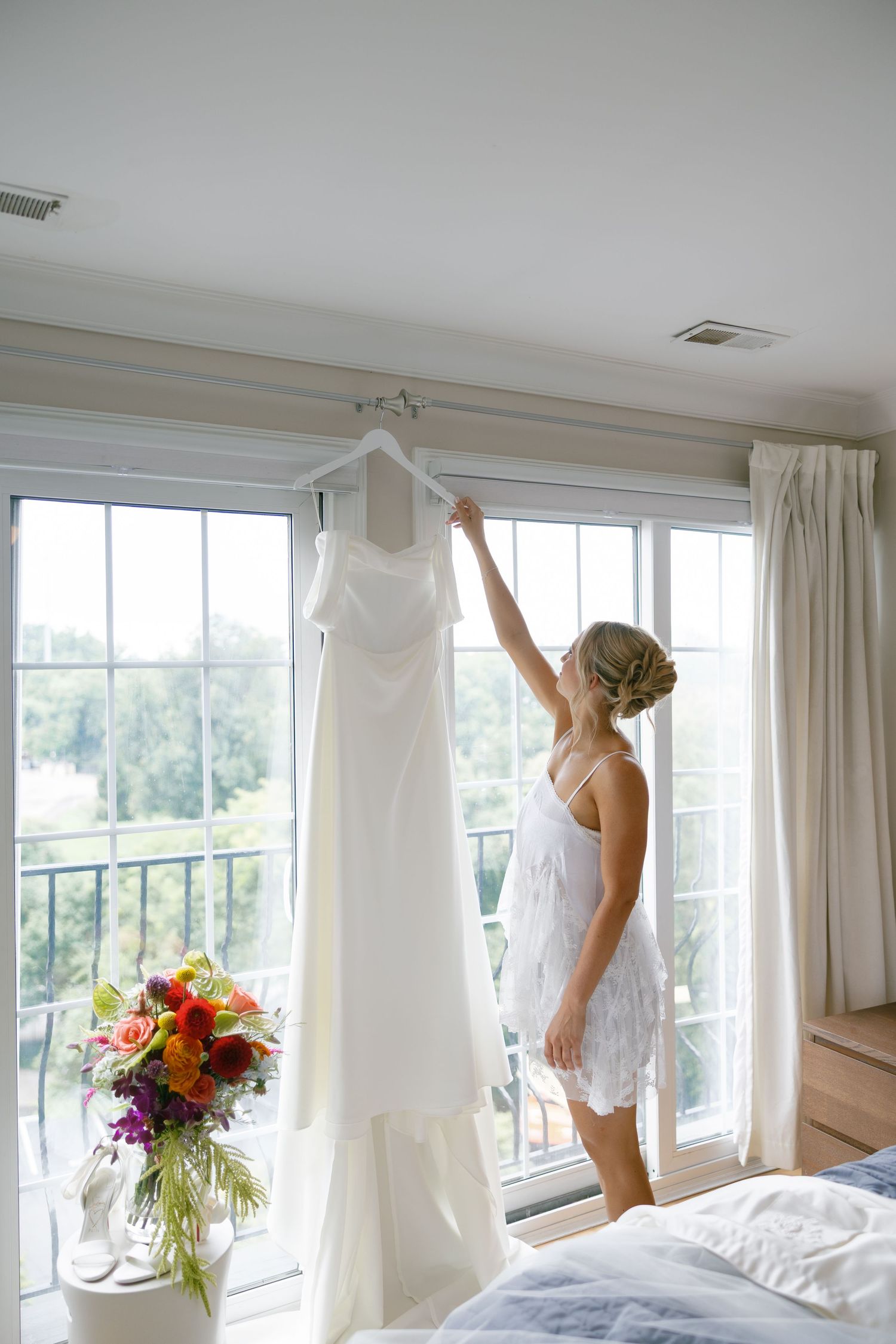 Bride in lace robe reaching up to hang white wedding dress on window in bright bedroom with colorful floral bouquet.