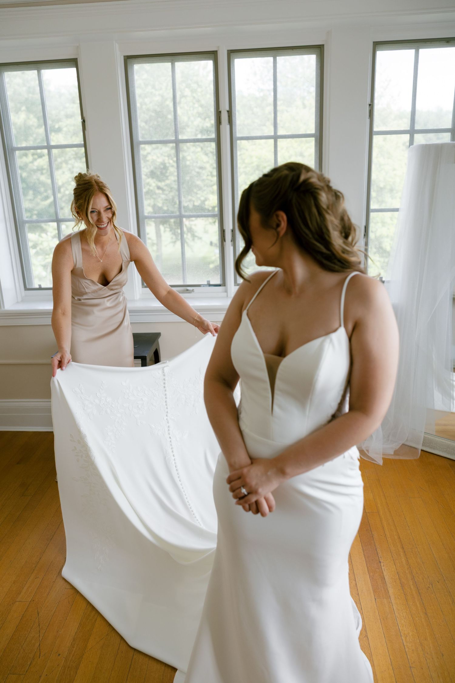 Bridesmaid helping the bride with her white wedding gown train in a bright sunlit room. French park wedding film