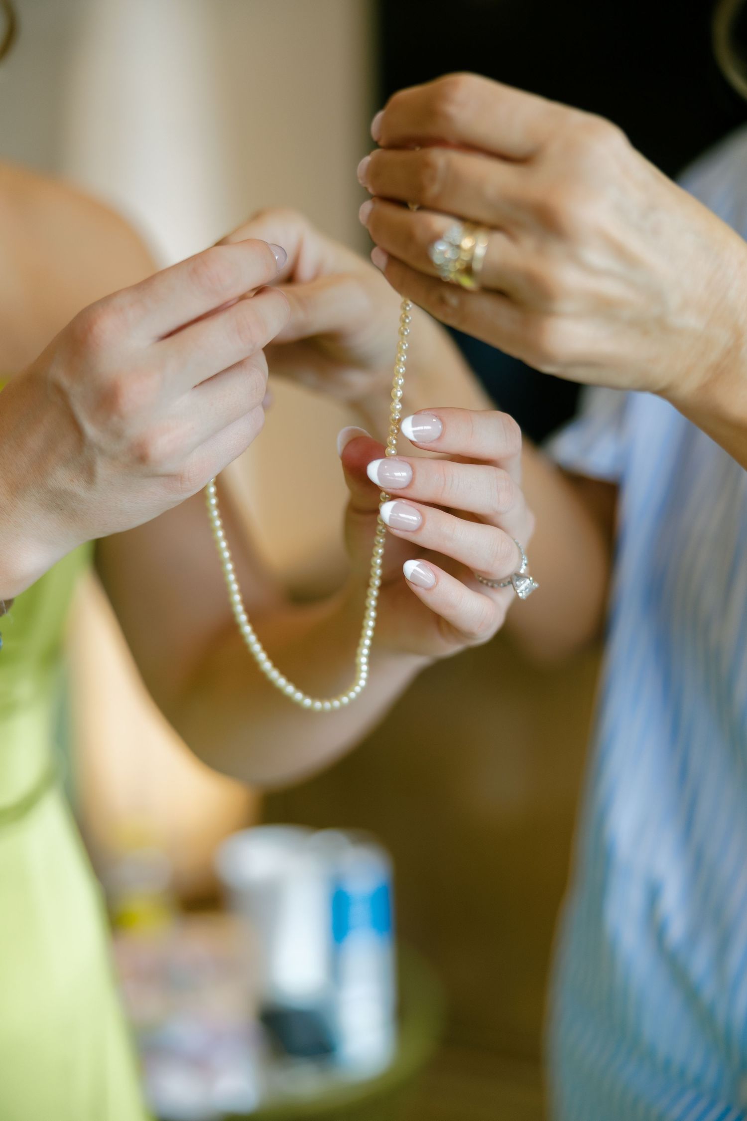 Two women's hands holding a delicate pearl necklace, one wearing a diamond ring with French manicure nails.