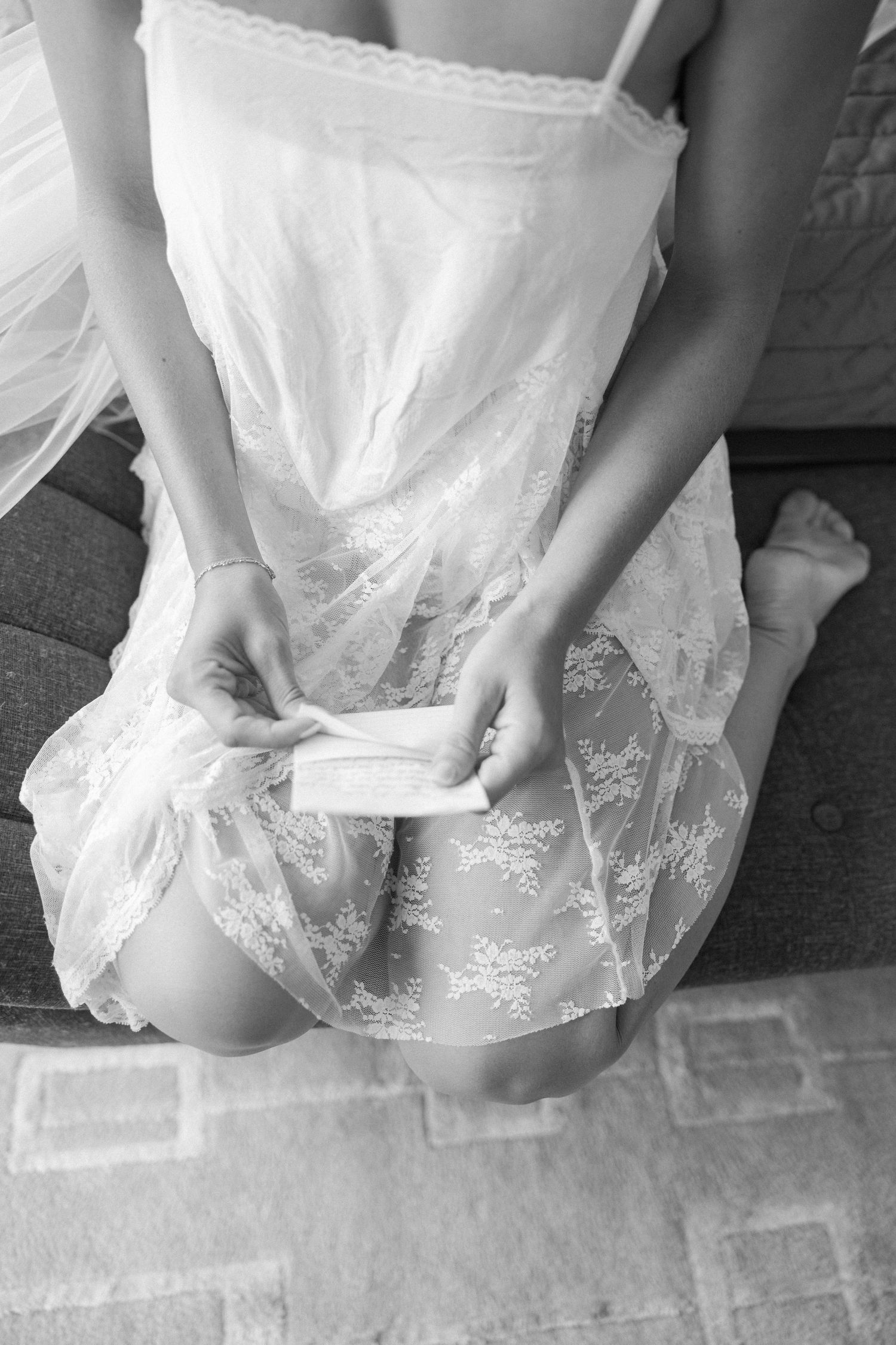 Black and white photo of a bride in lace dress reading a letter while seated on a couch before her wedding.