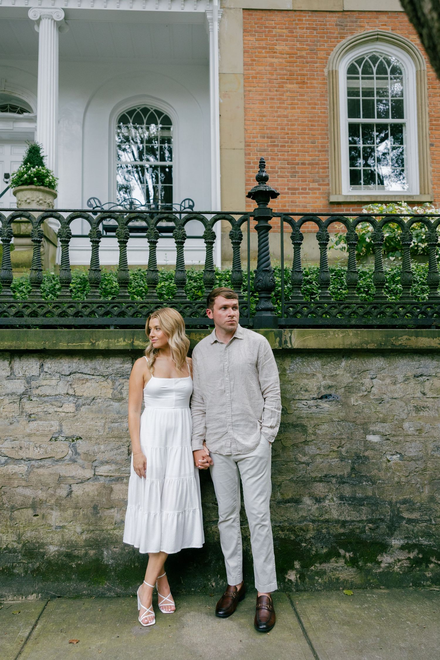 Couple posing together in front of historic brick building with ornate iron fence on stone wall.