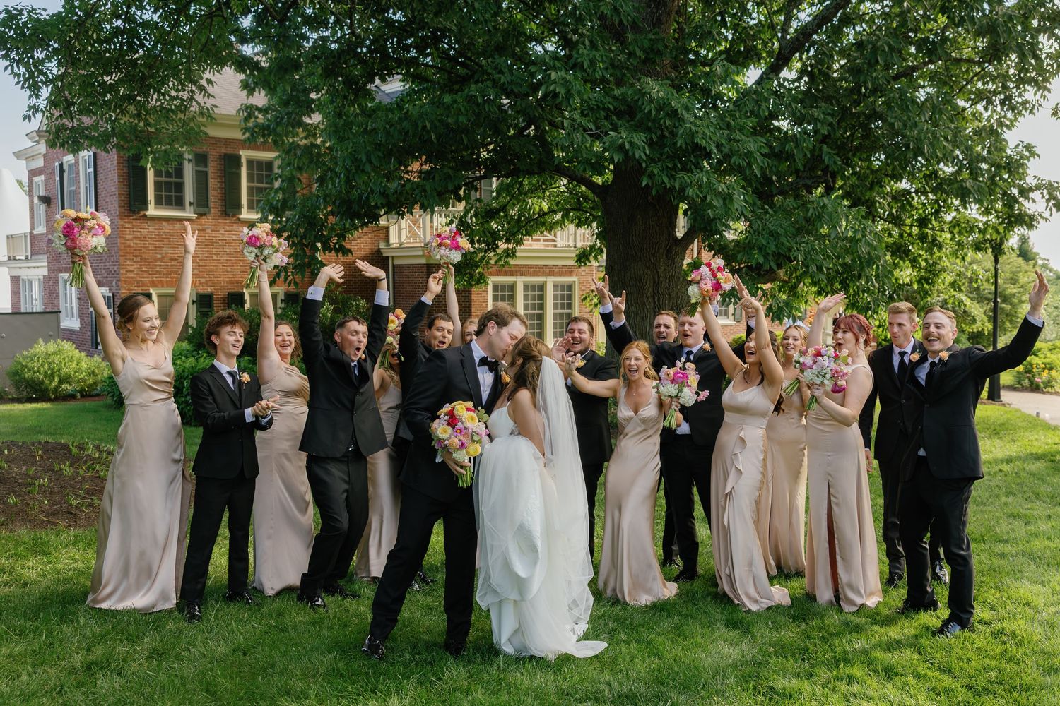 Joyful wedding party cheers under a large tree with bridesmaids in champagne gowns and groomsmen in black suits.