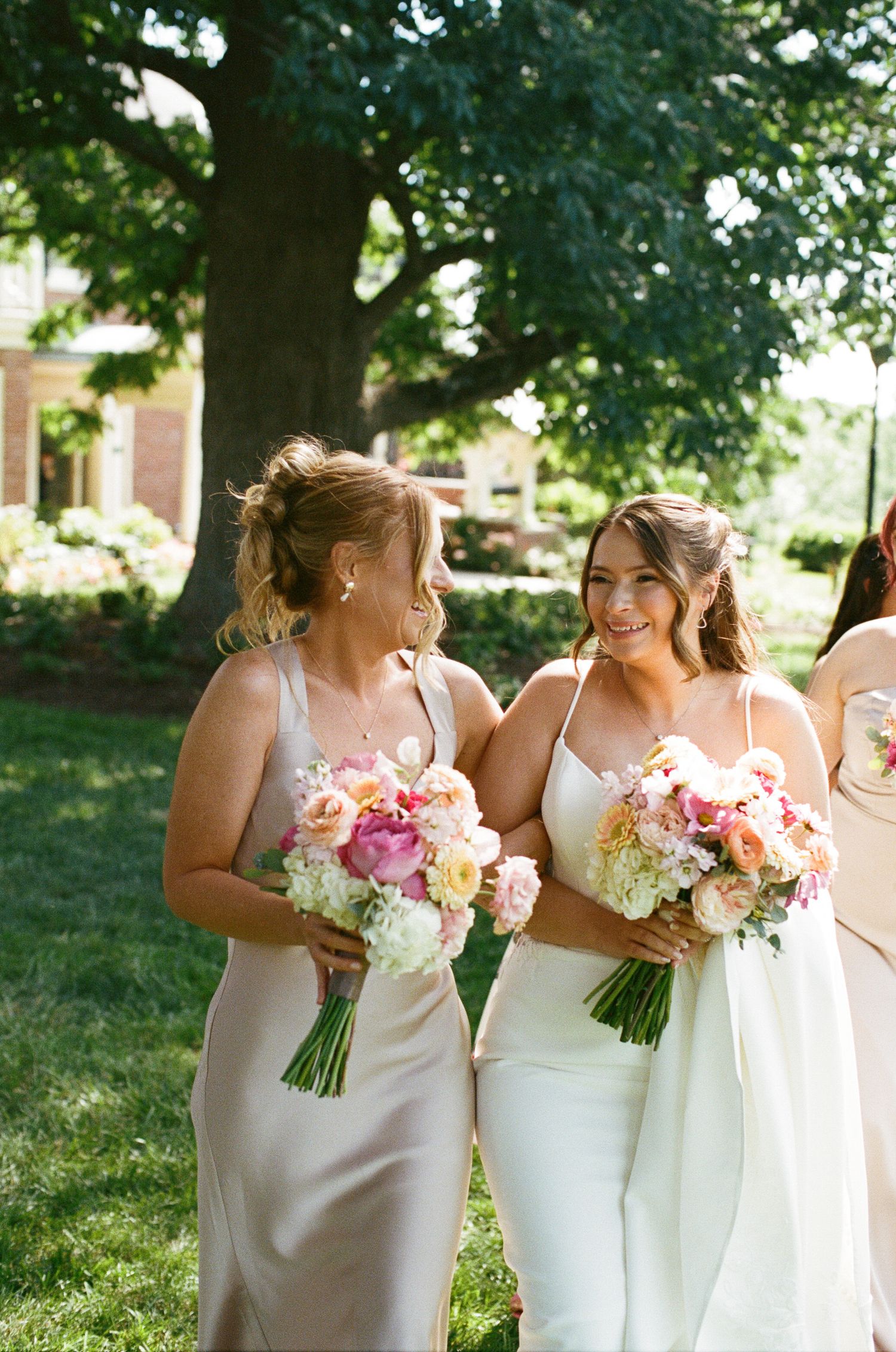 Two bridesmaids in champagne and white gowns laughing while holding pink and white floral bouquets outdoors.