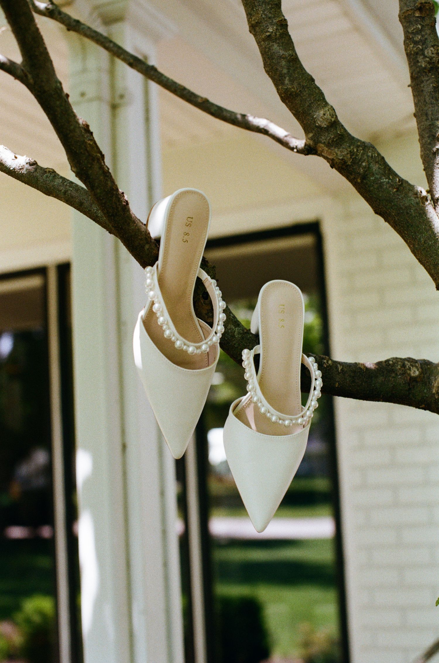 Elegant white pointed-toe bridal heels with pearl ankle straps displayed on tree branch outdoors.