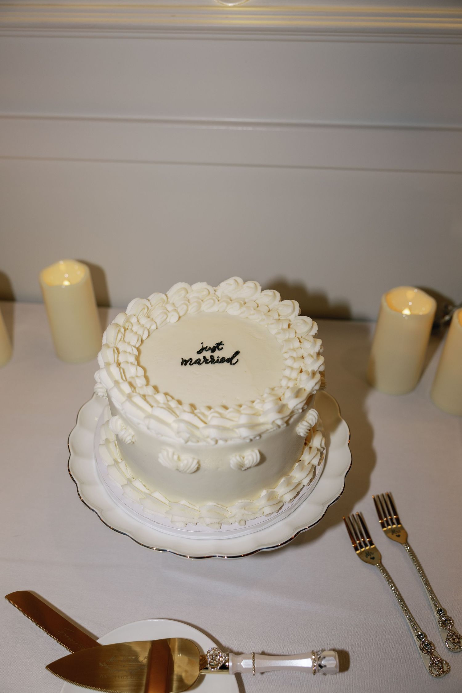 Elegant two-tier white wedding cake inscribed with just married surrounded by pillar candles on a white table.