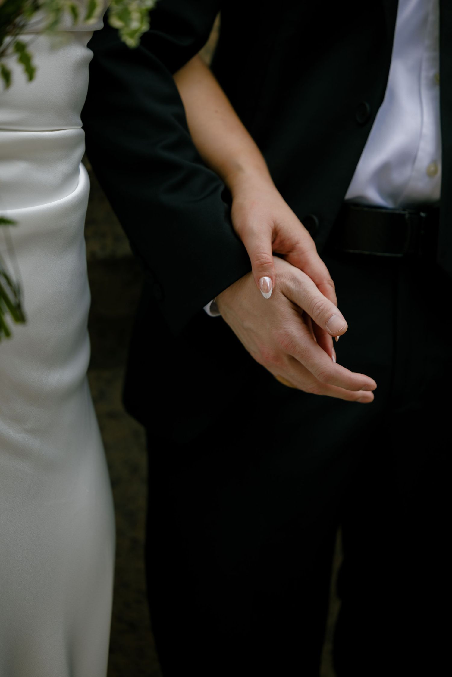 Close-up of bride and groom gently holding hands, bride in white satin gown with manicured nails.