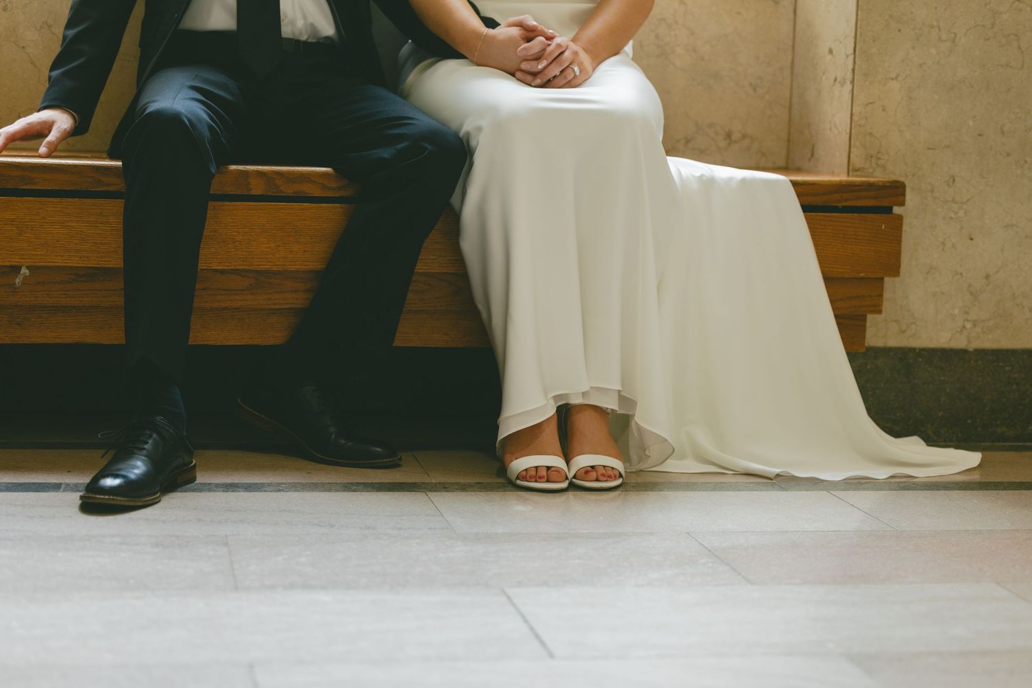Close-up of bride in white dress and groom in suit sitting together on wooden bench.