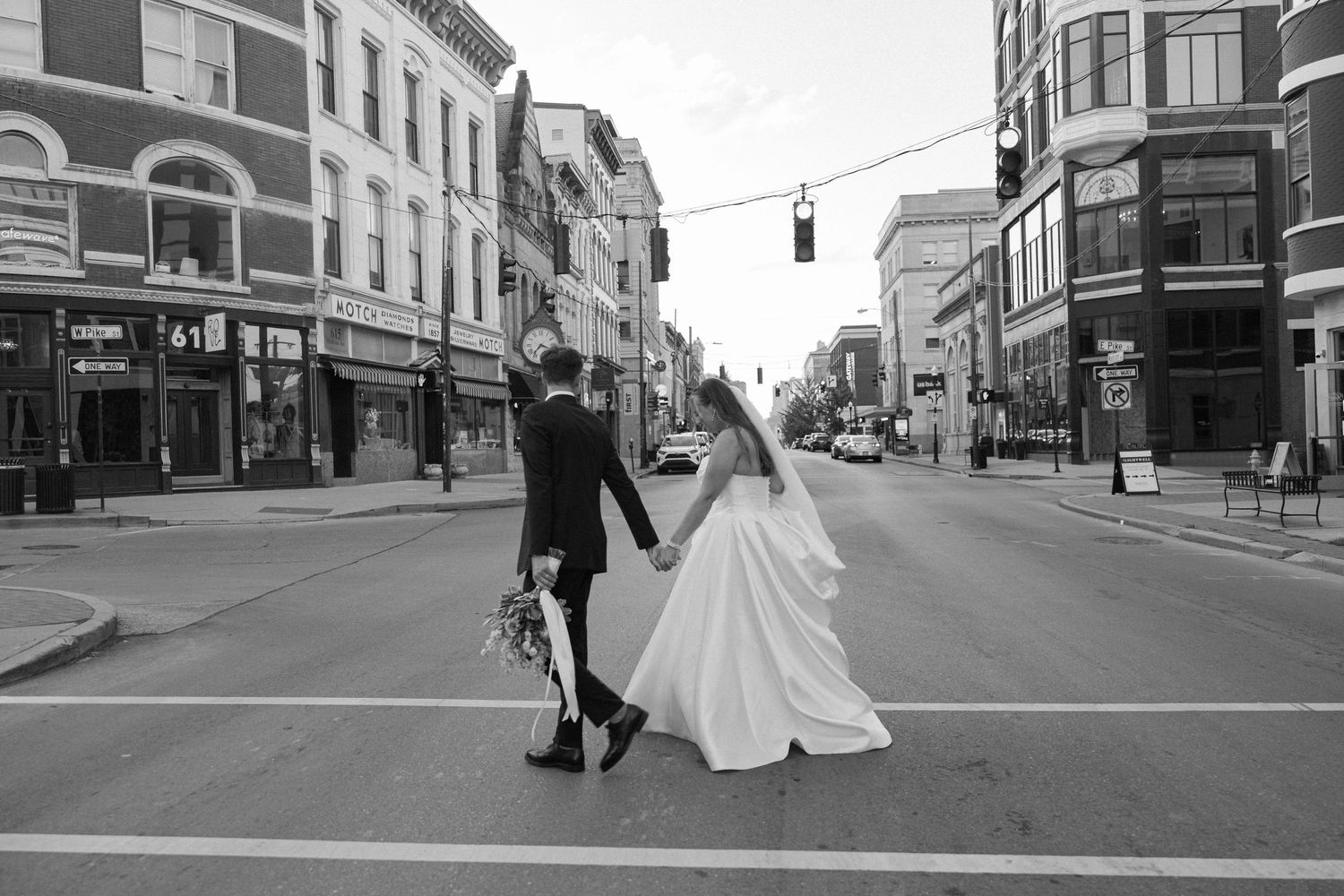 Black and white photo of bride and groom holding hands while crossing a city street, bride's gown flowing behind her.